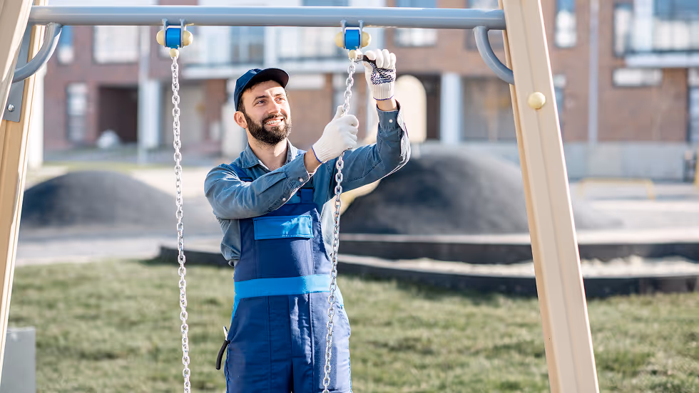 A man in blue overalls installs a playground swing, smiling. He wears a cap and gloves. Background: grassy area, blurred buildings, sunny day.