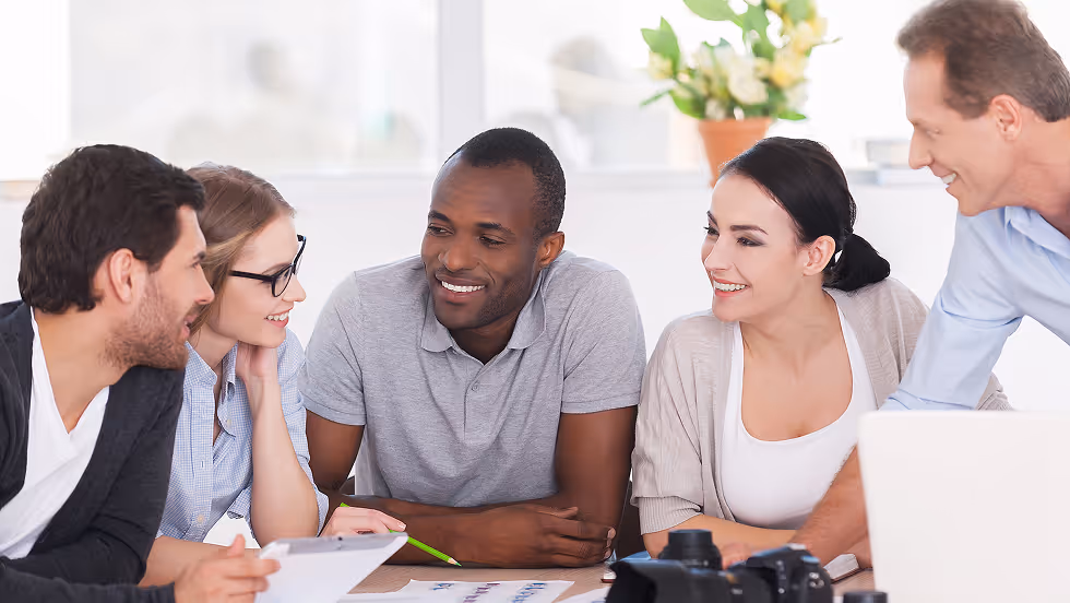 A diverse group of five people in a casual meeting setting, smiling and engaged in discussion around a table with papers, creating a collaborative atmosphere.