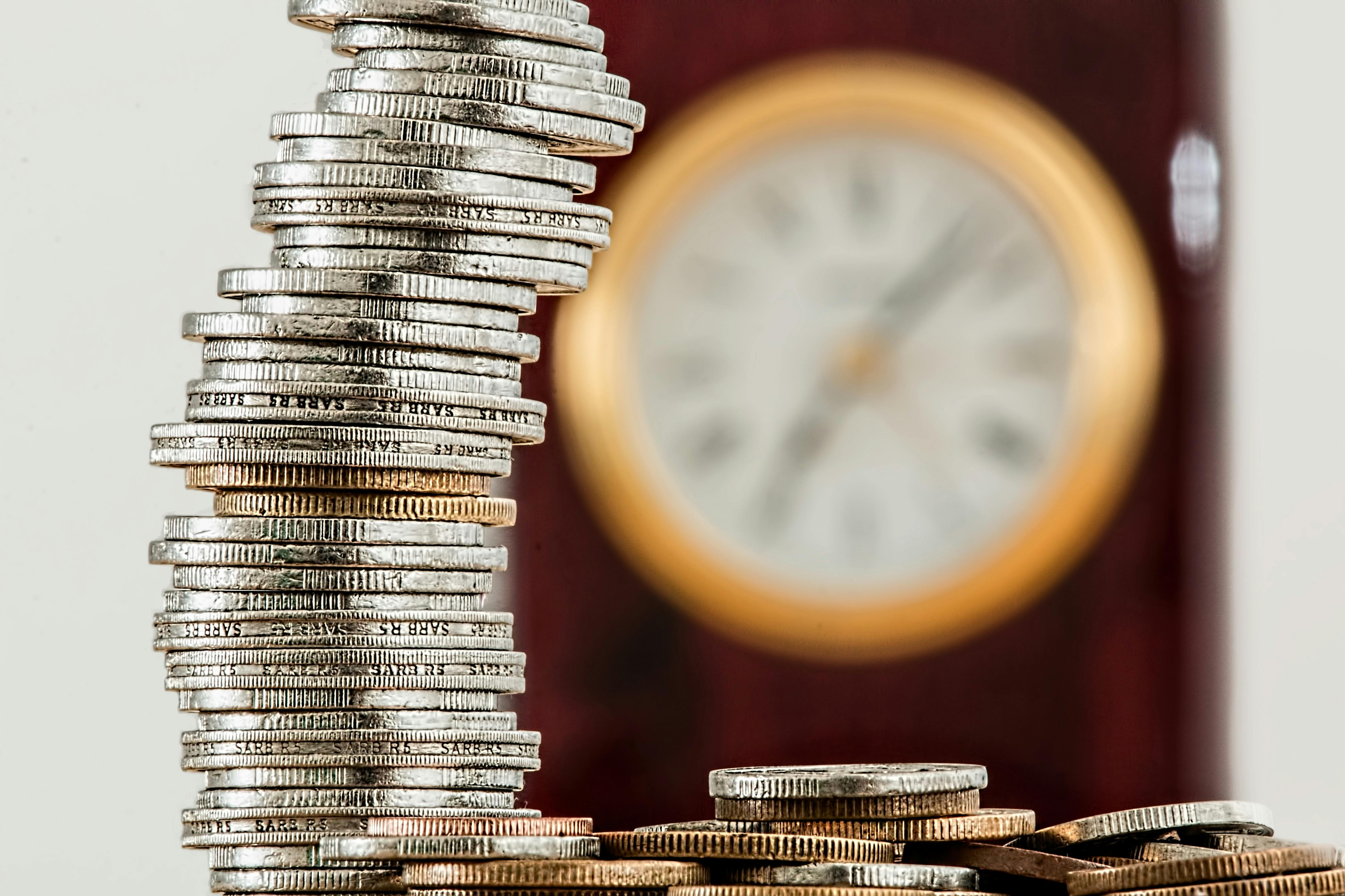 Stack of coins in the foreground with a blurred clock in the background, suggesting saving money over time.