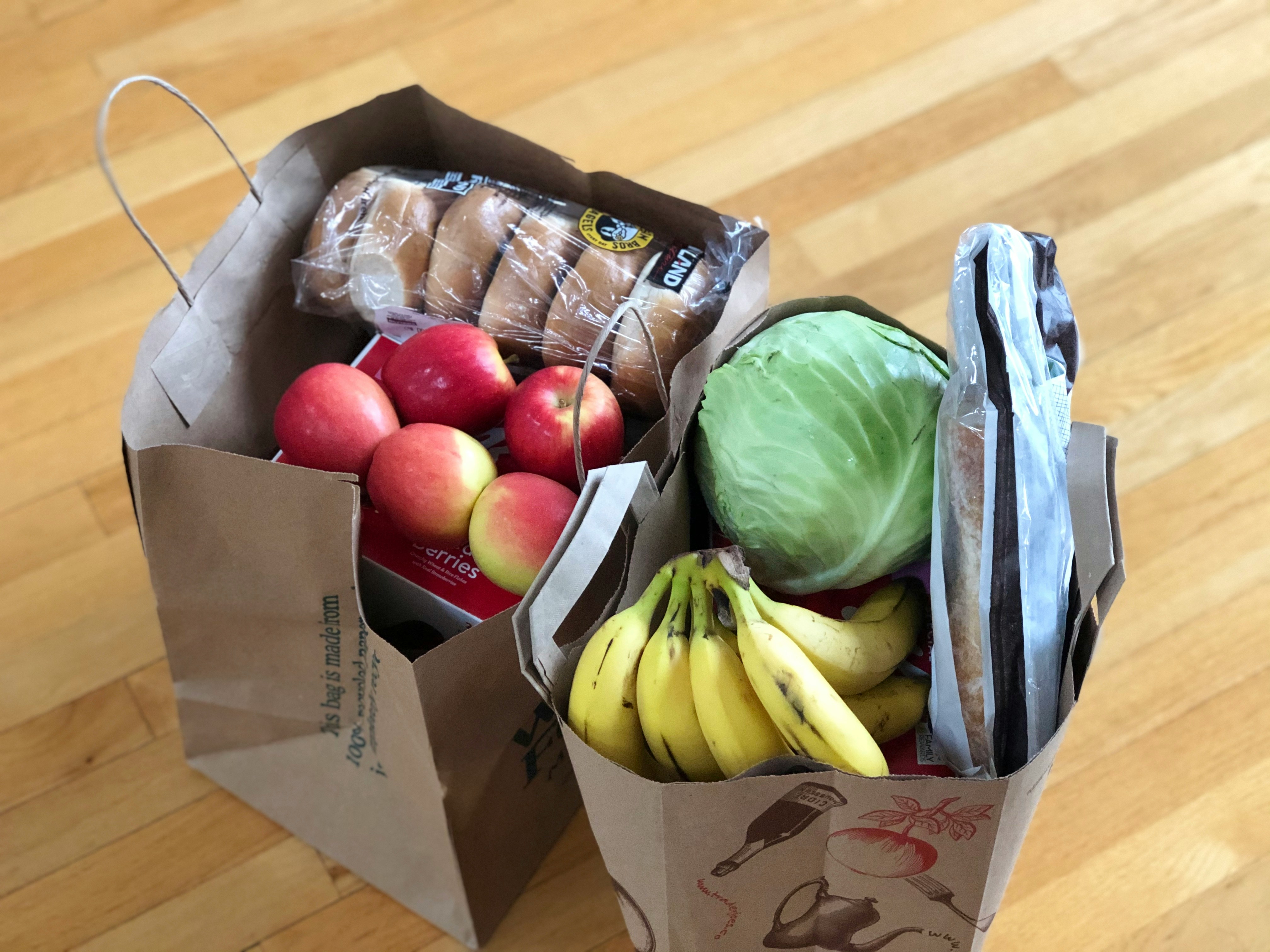 Reusable grocery bags filled with apples, bananas, cabbage, and packaged bread on a wooden floor.