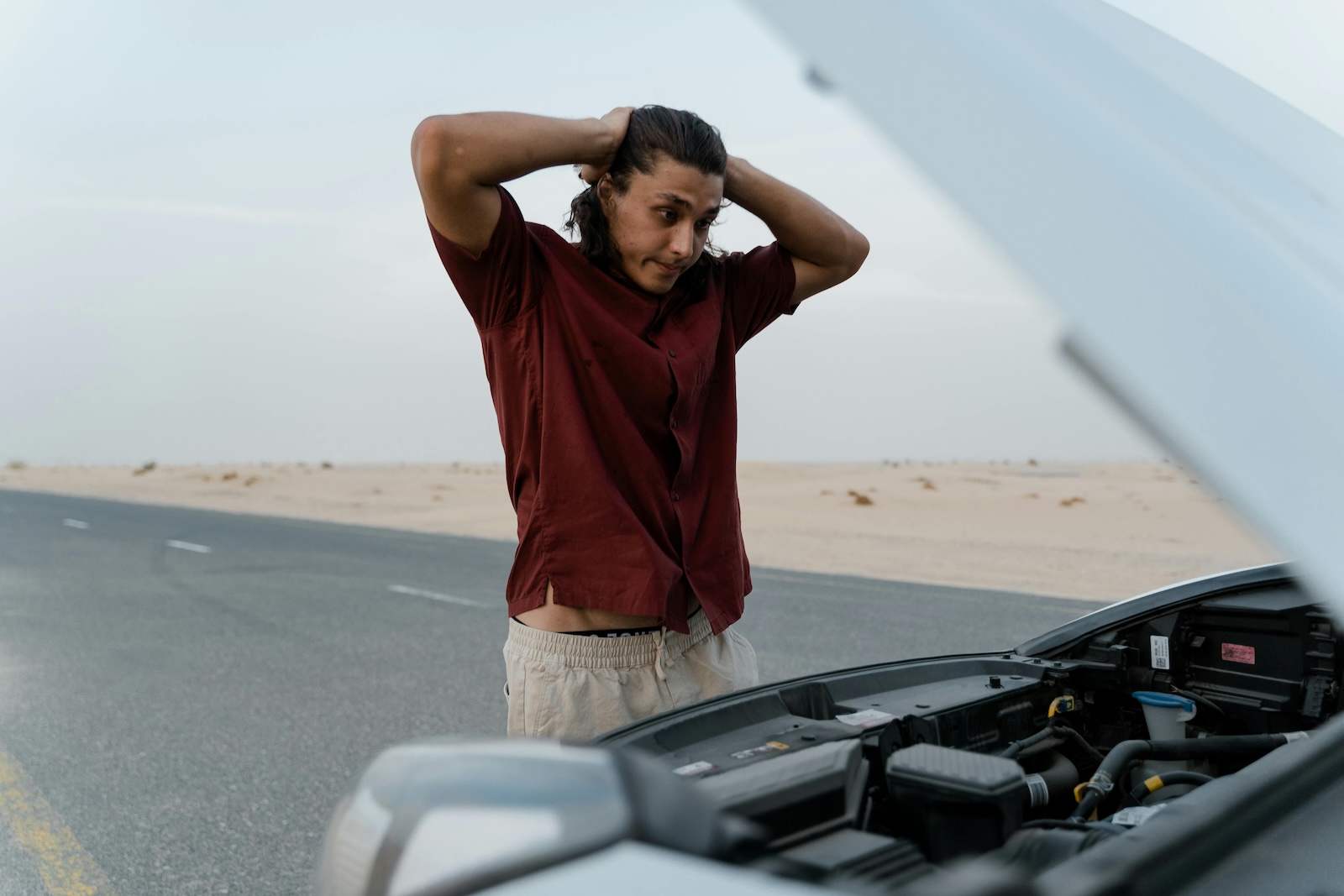 Person standing on the roadside with hands on their head while looking into an open car hood in a desert setting.