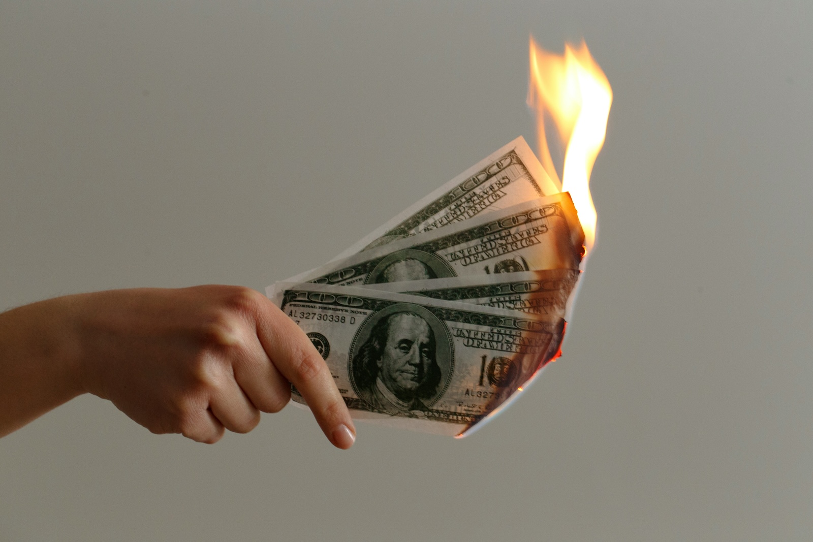 Hand holding U.S. dollar bills that are on fire, symbolizing money loss or high financial costs.