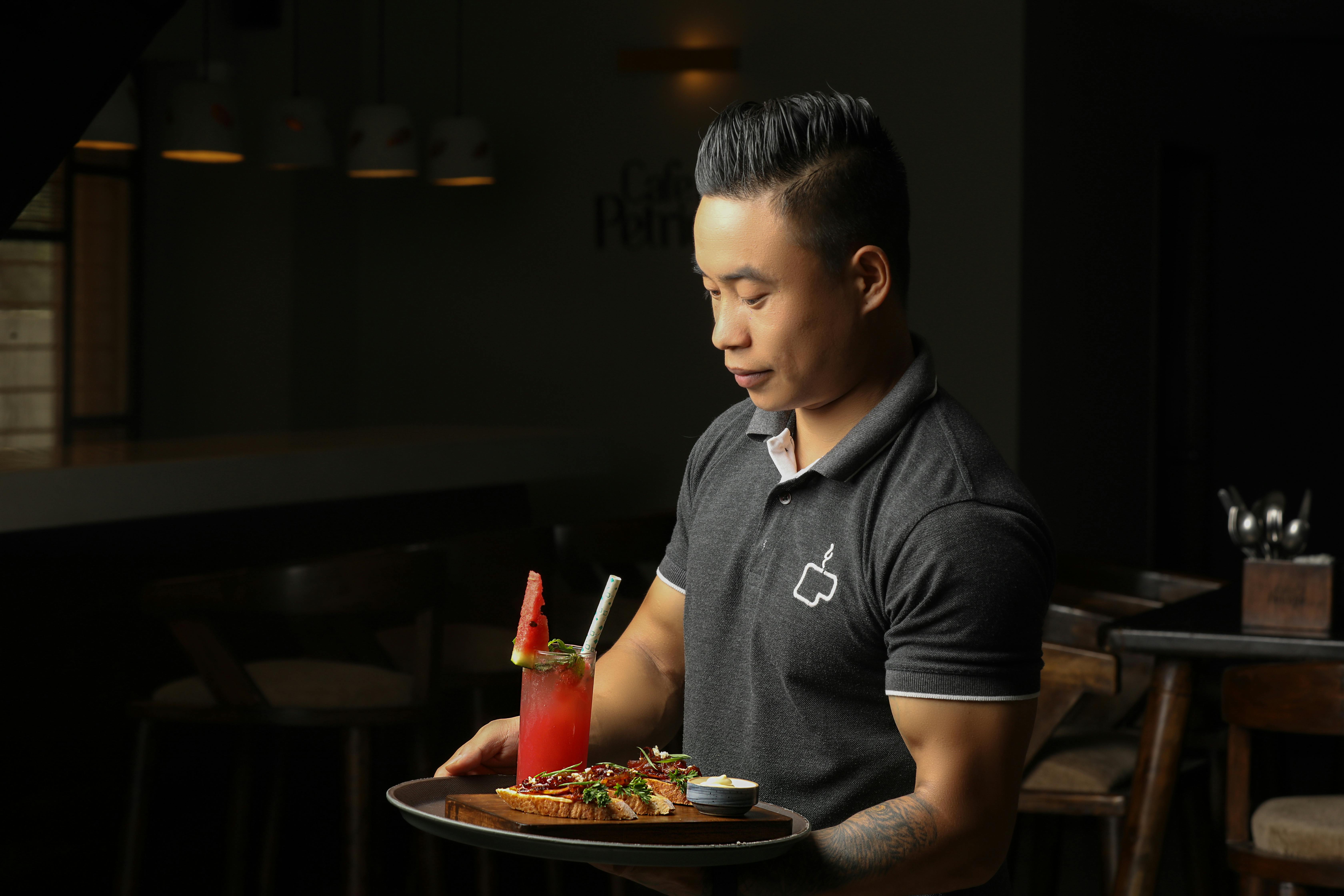 Server carrying a tray with food and a watermelon drink inside a dimly lit restaurant.