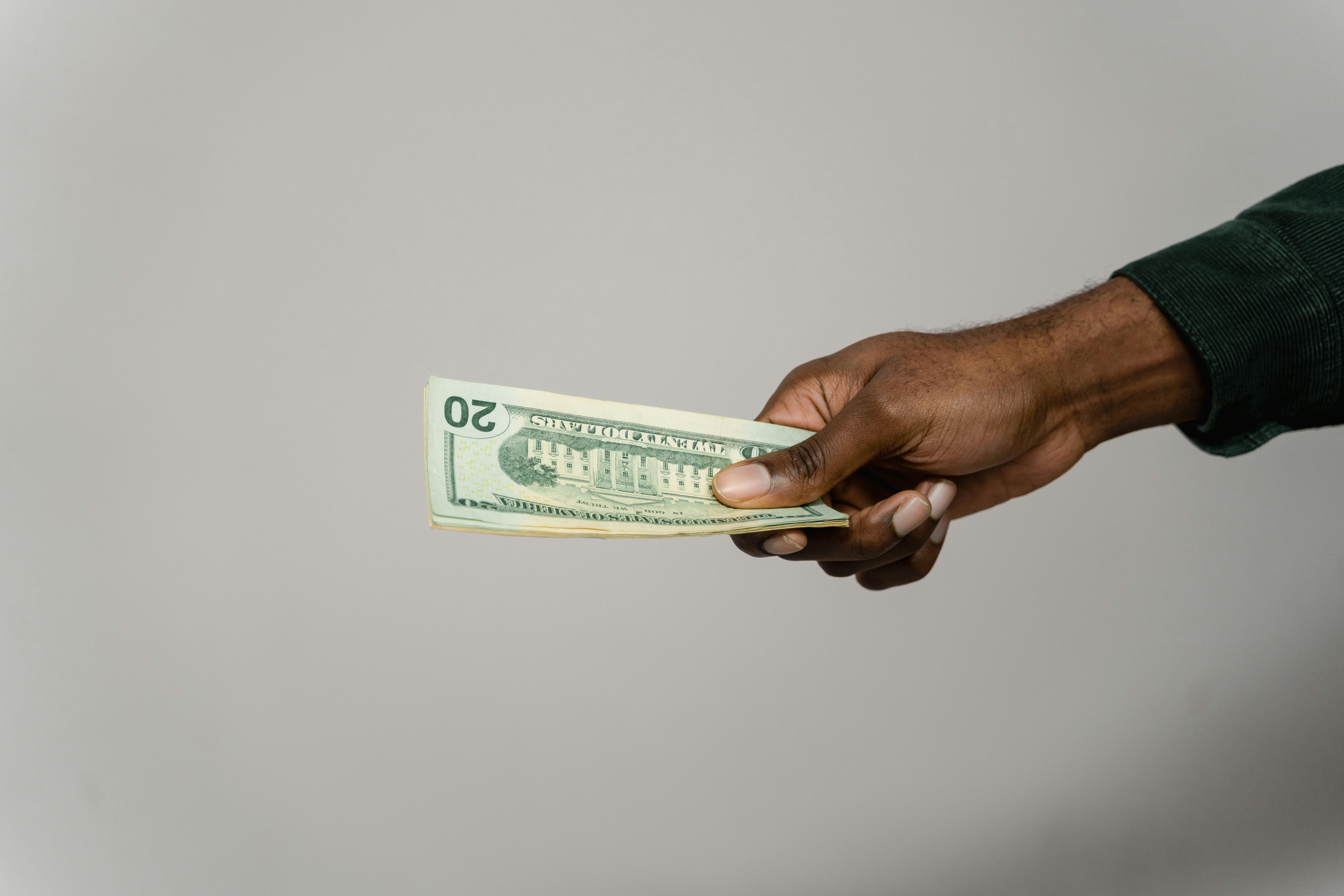 A hand extending a stack of $20 bills against a light gray background.