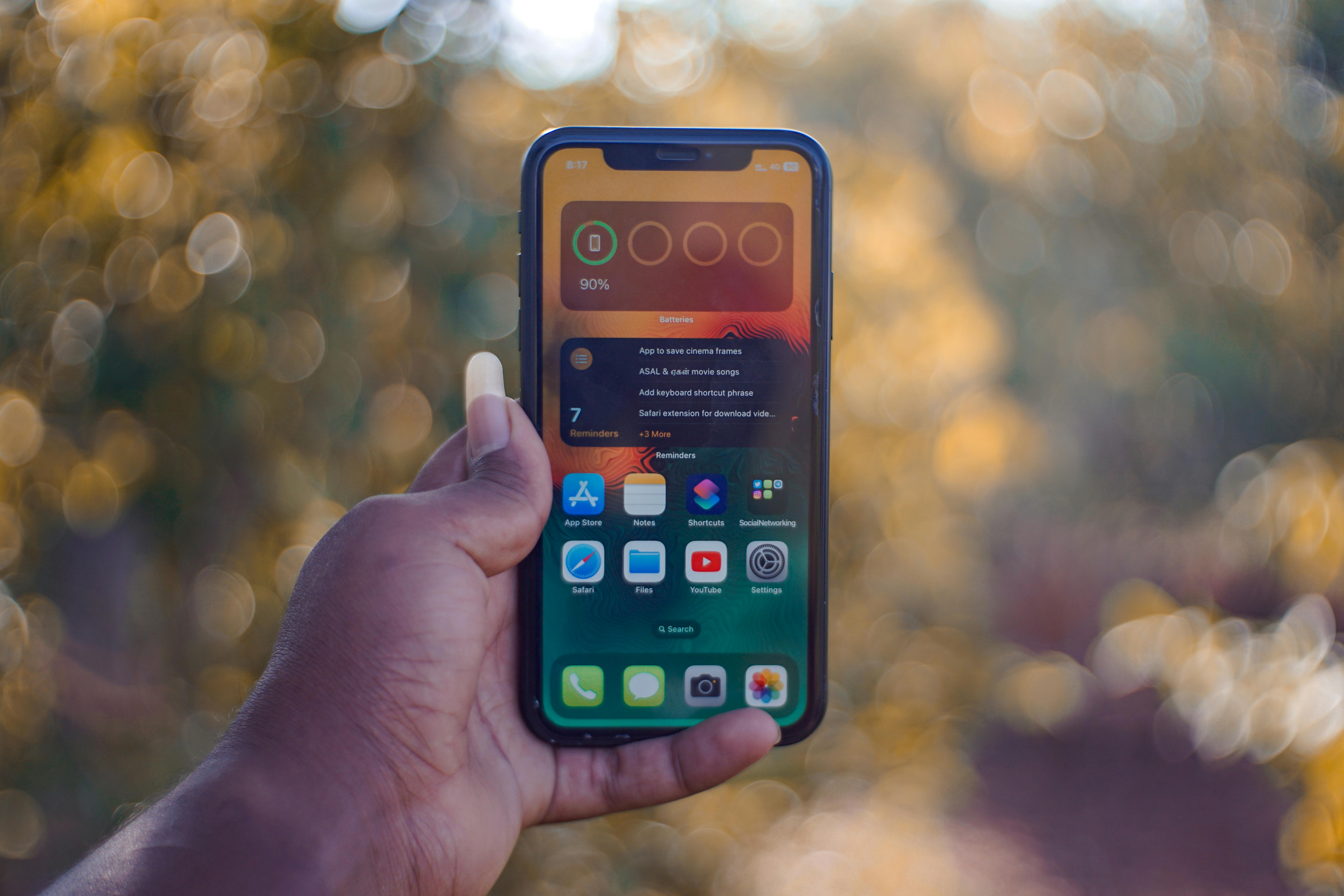 A hand holding an iPhone displaying its home screen with widgets, set against a blurred outdoor background.