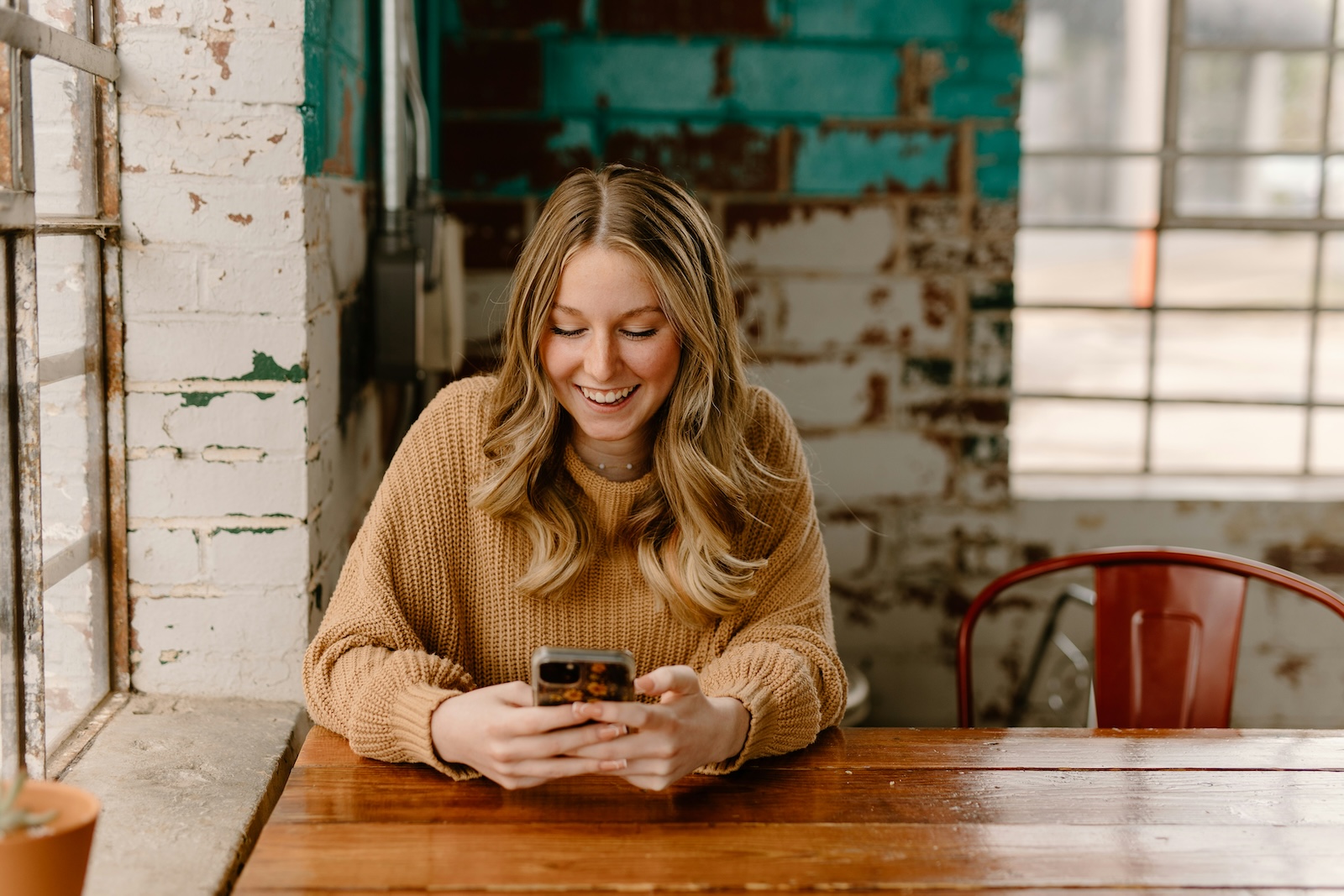 A young woman smiles while looking at her phone at a wooden table in a rustic café.