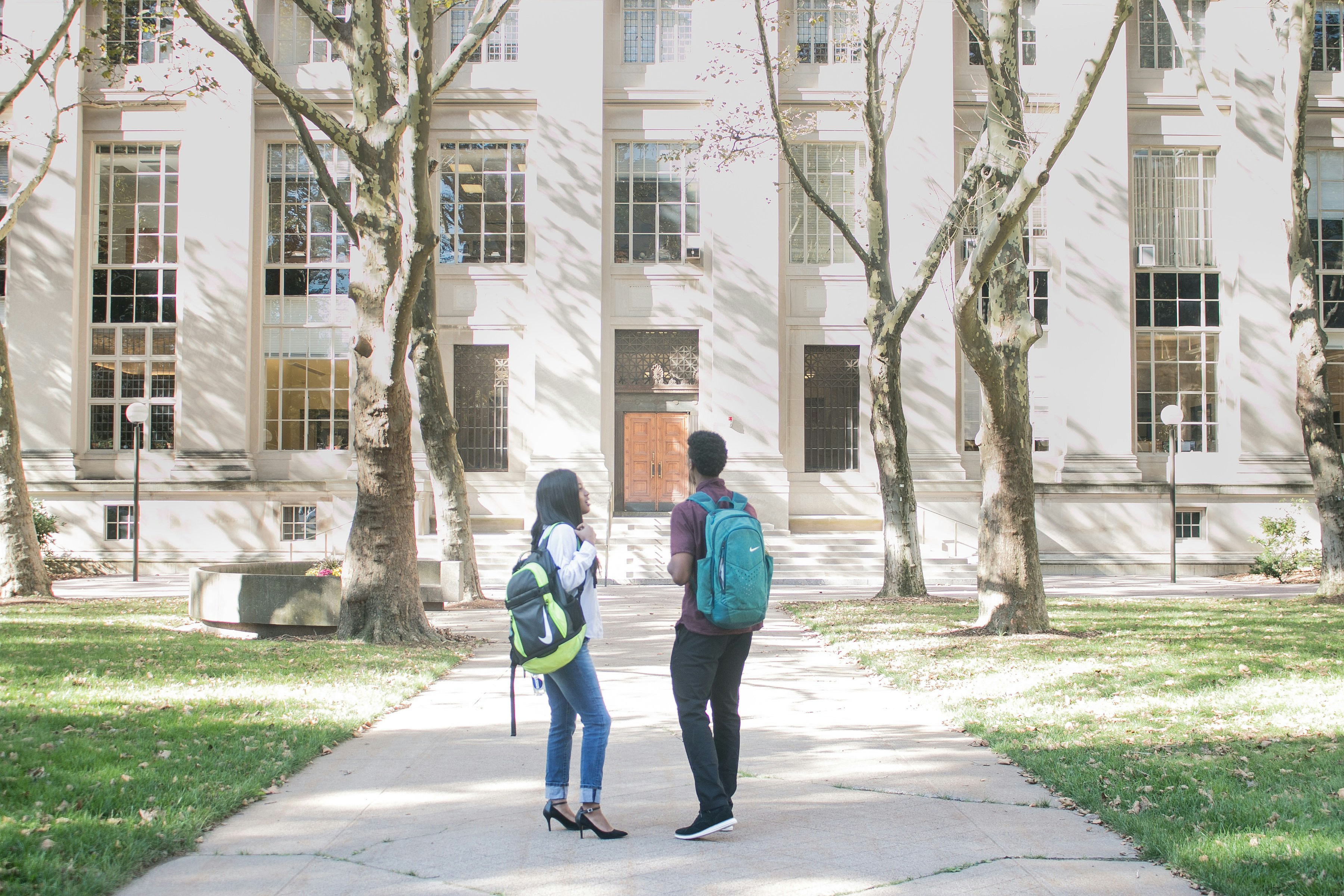 Two college students wearing backpacks chat on a sunny campus walkway in front of a large academic building.