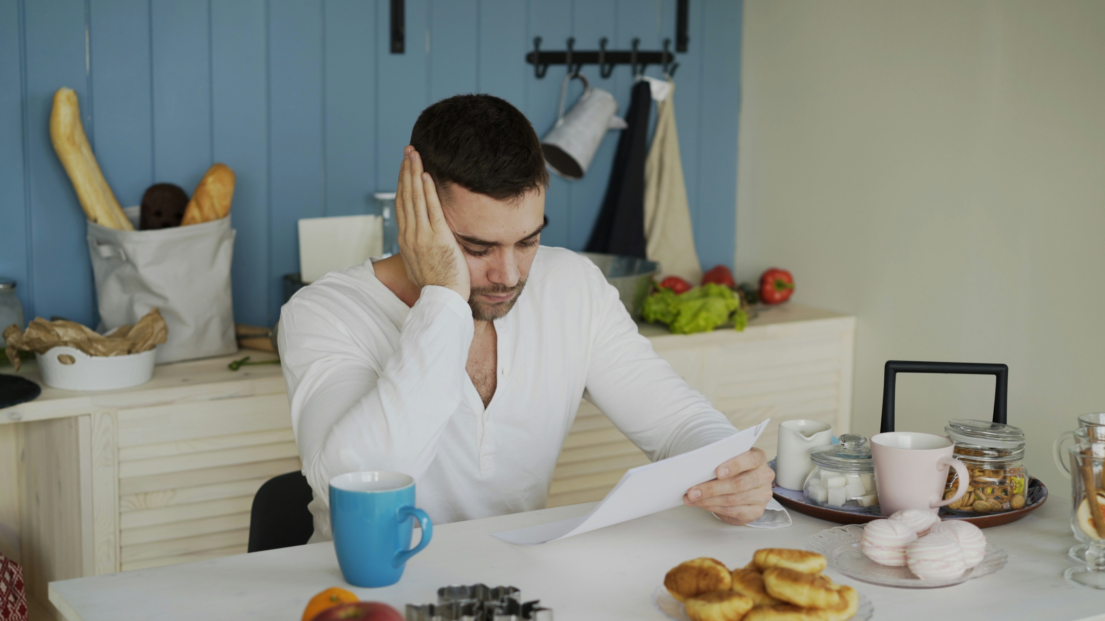 A stressed man sitting at a kitchen table reading a document with his hand on his head.