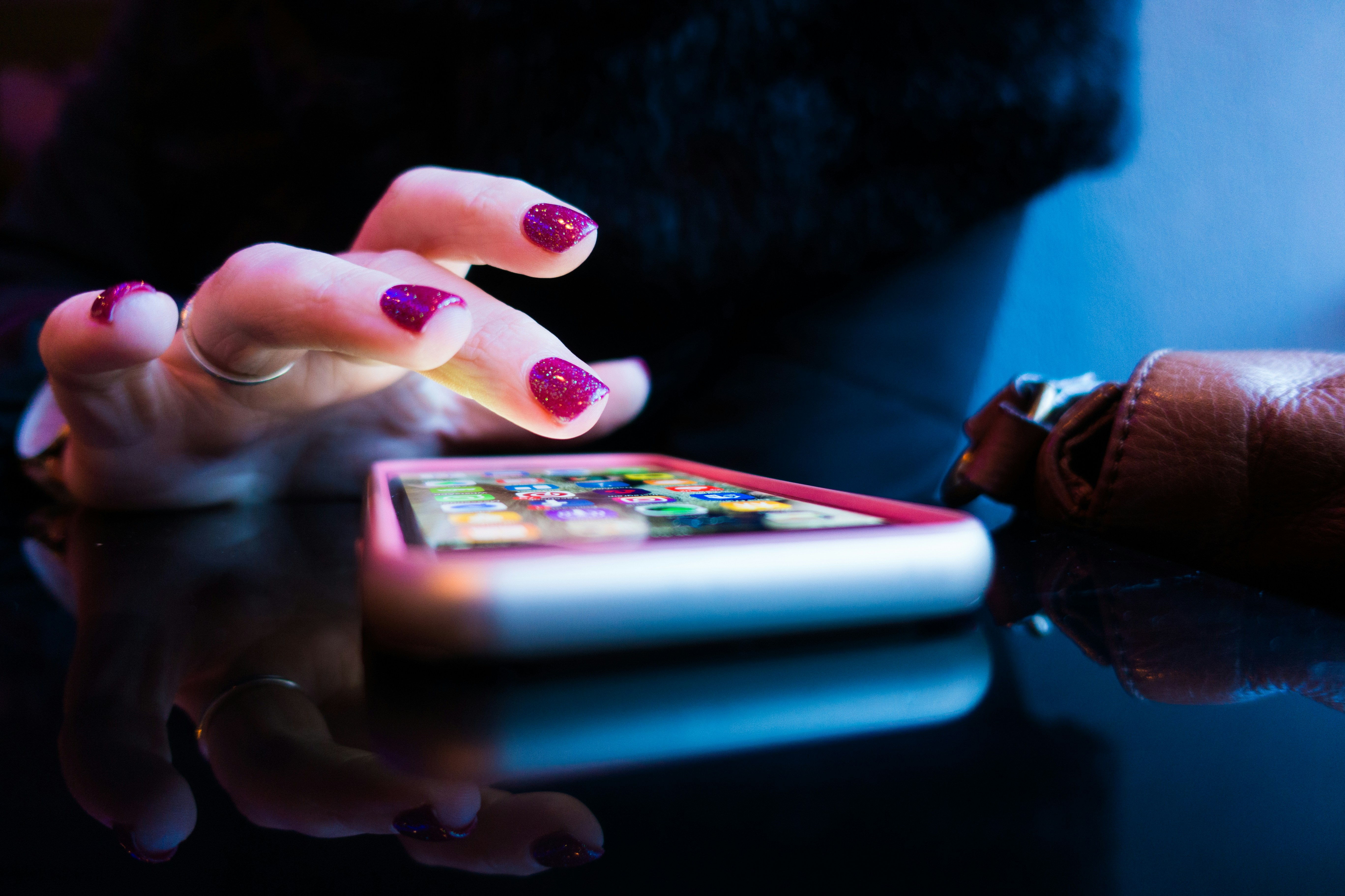 A close-up of a hand with glittery pink nails reaching toward a smartphone displaying a home screen of colorful apps.