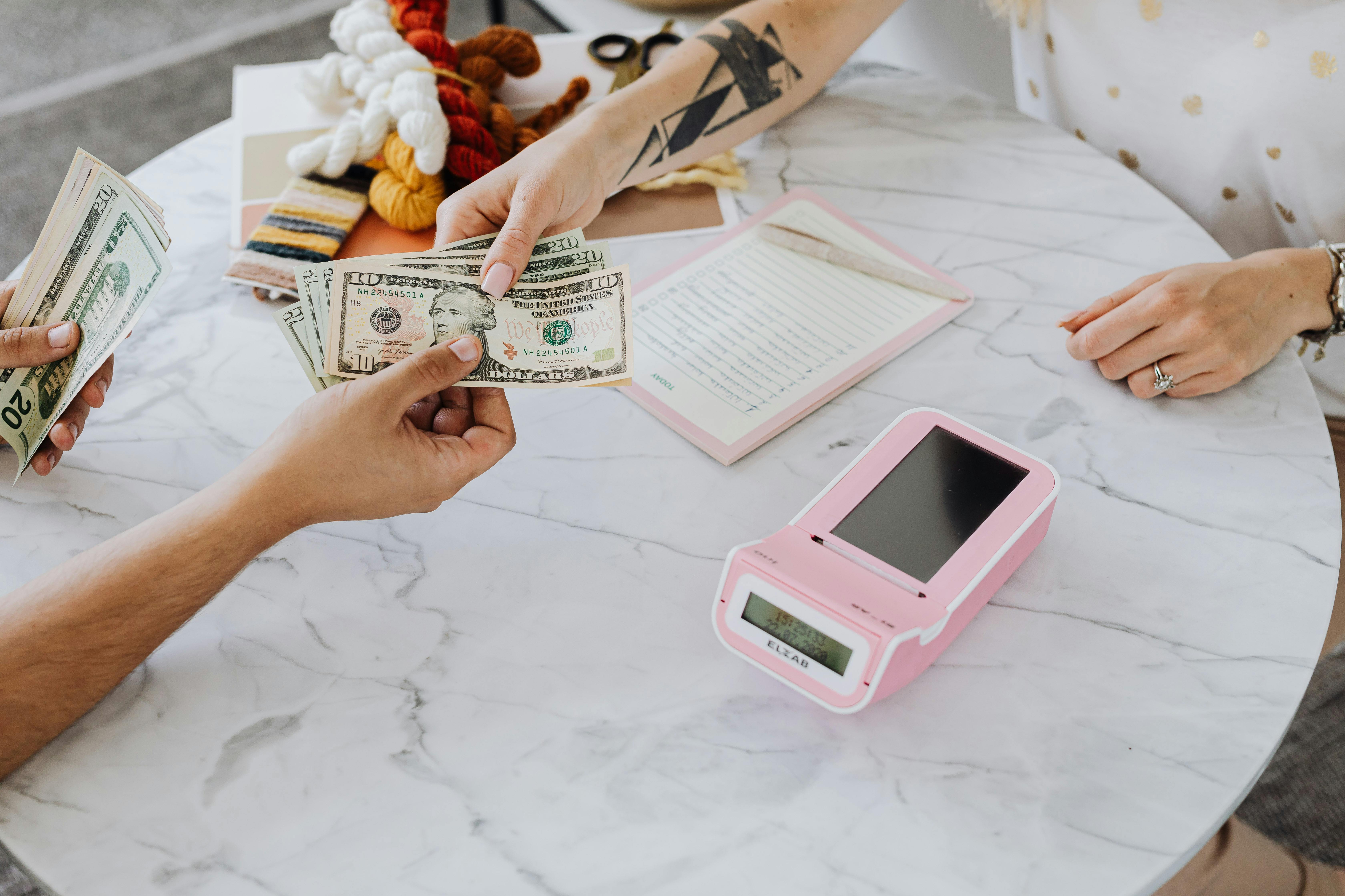 Claude responded: Two people exchange cash across a marble table, with a pink card reader and open notebook nearby.Two people exchange cash across a marble table, with a pink card reader and open notebook nearby.