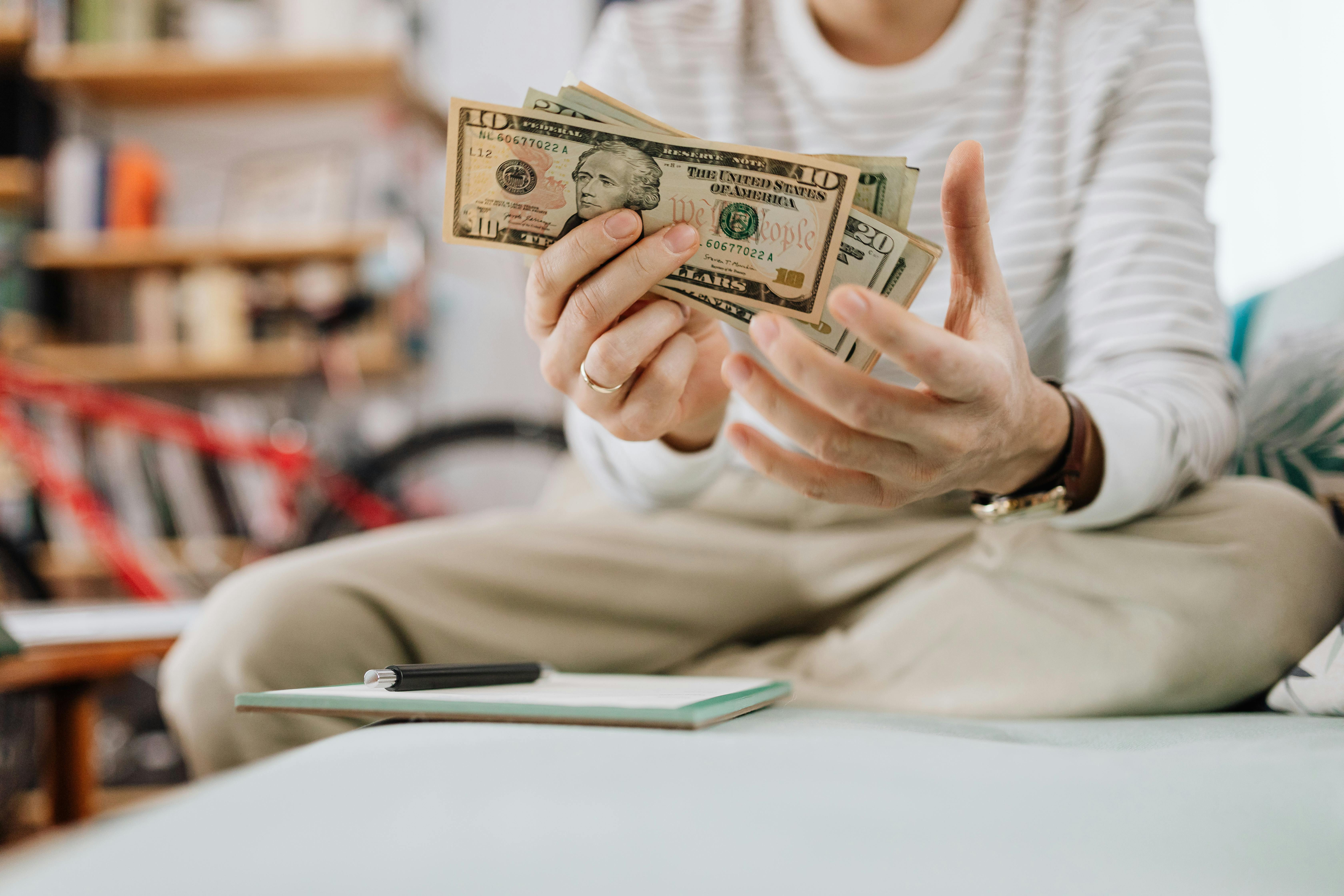 Claude responded: A person counting a handful of US dollar bills, with a notebook and pen on the table in front of them.A person counting a handful of US dollar bills, with a notebook and pen on the table in front of them.