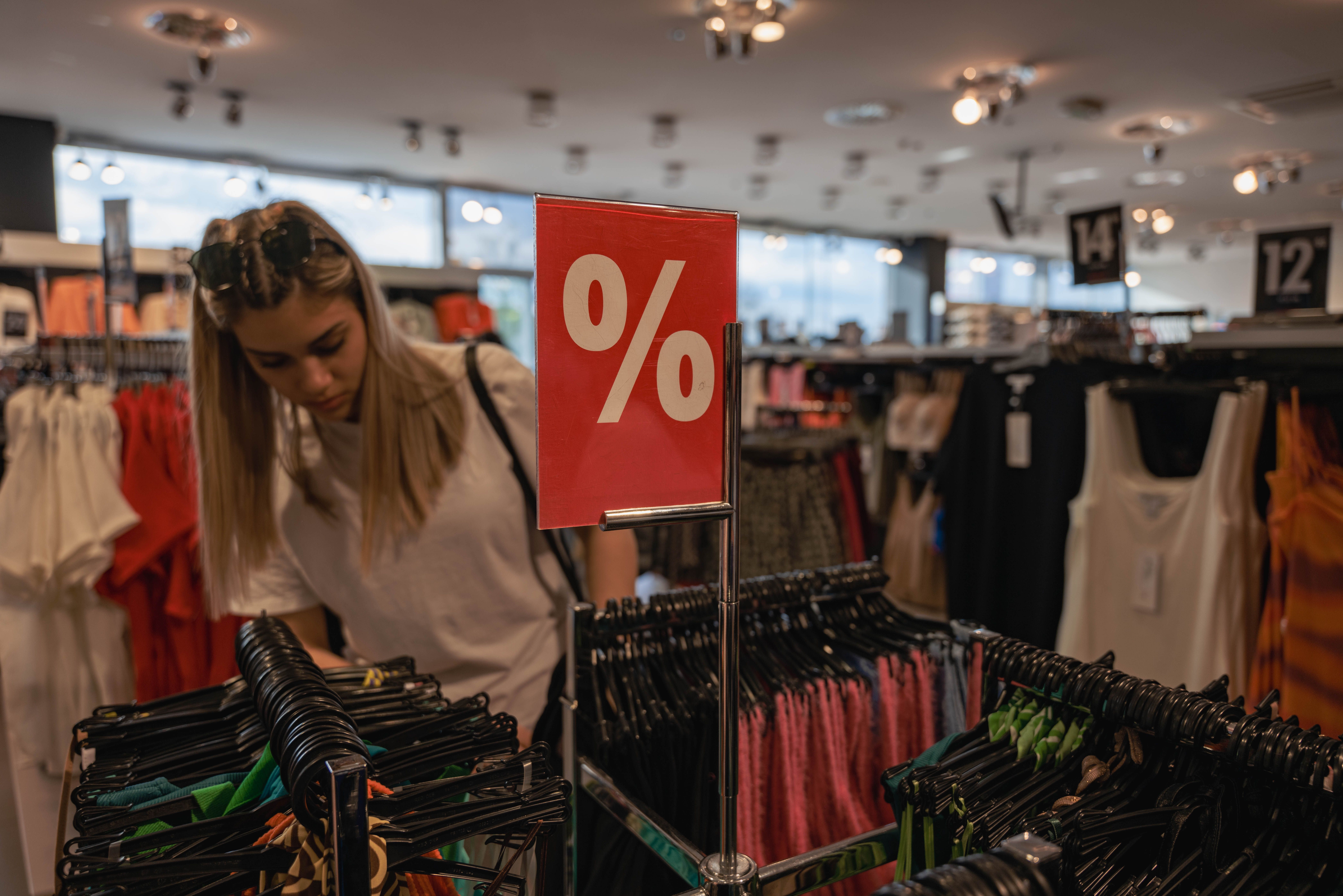 A shopper browses clothing racks near a red percent-off discount sign in a retail store.