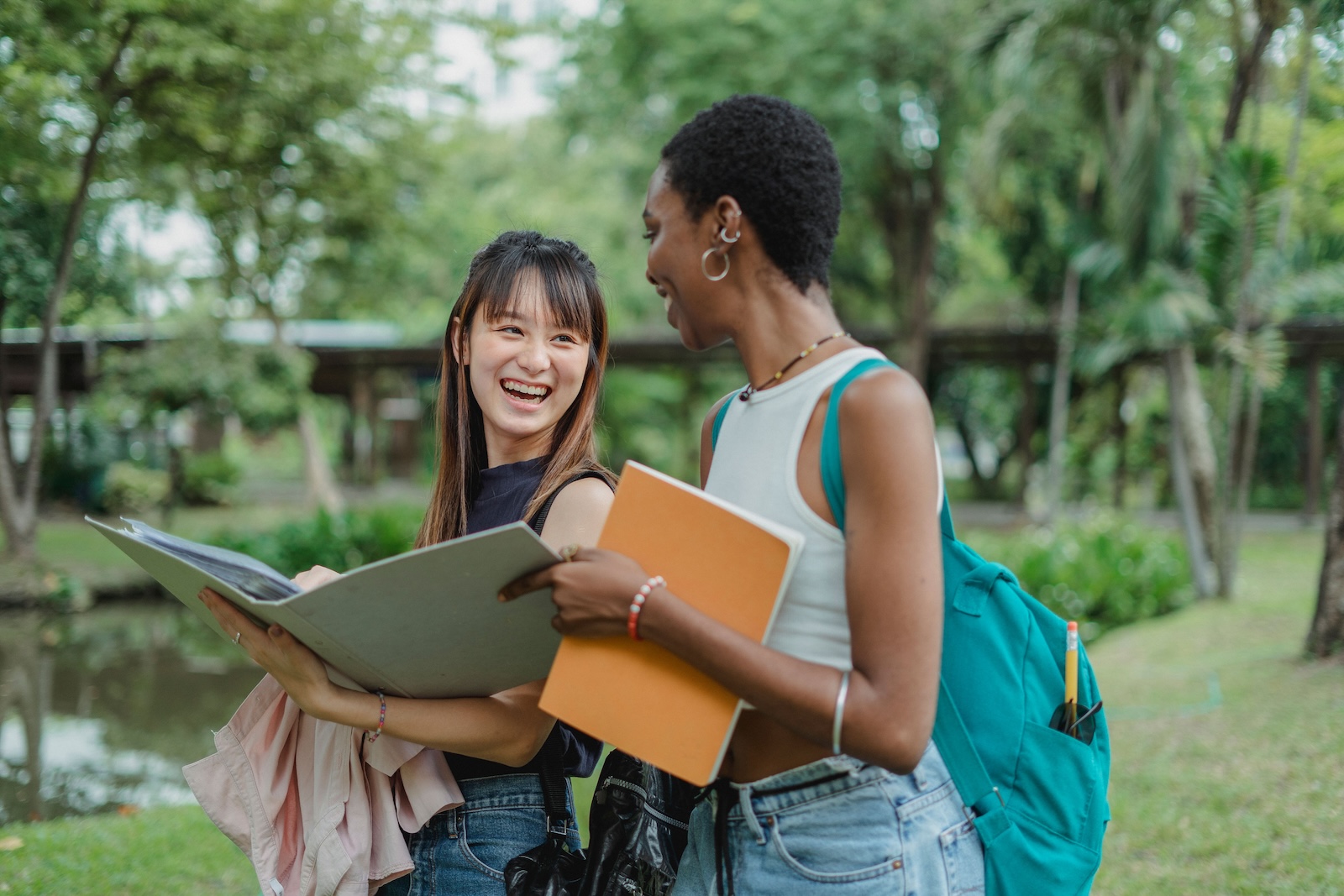 Two students carrying notebooks and backpacks laugh together in a park.