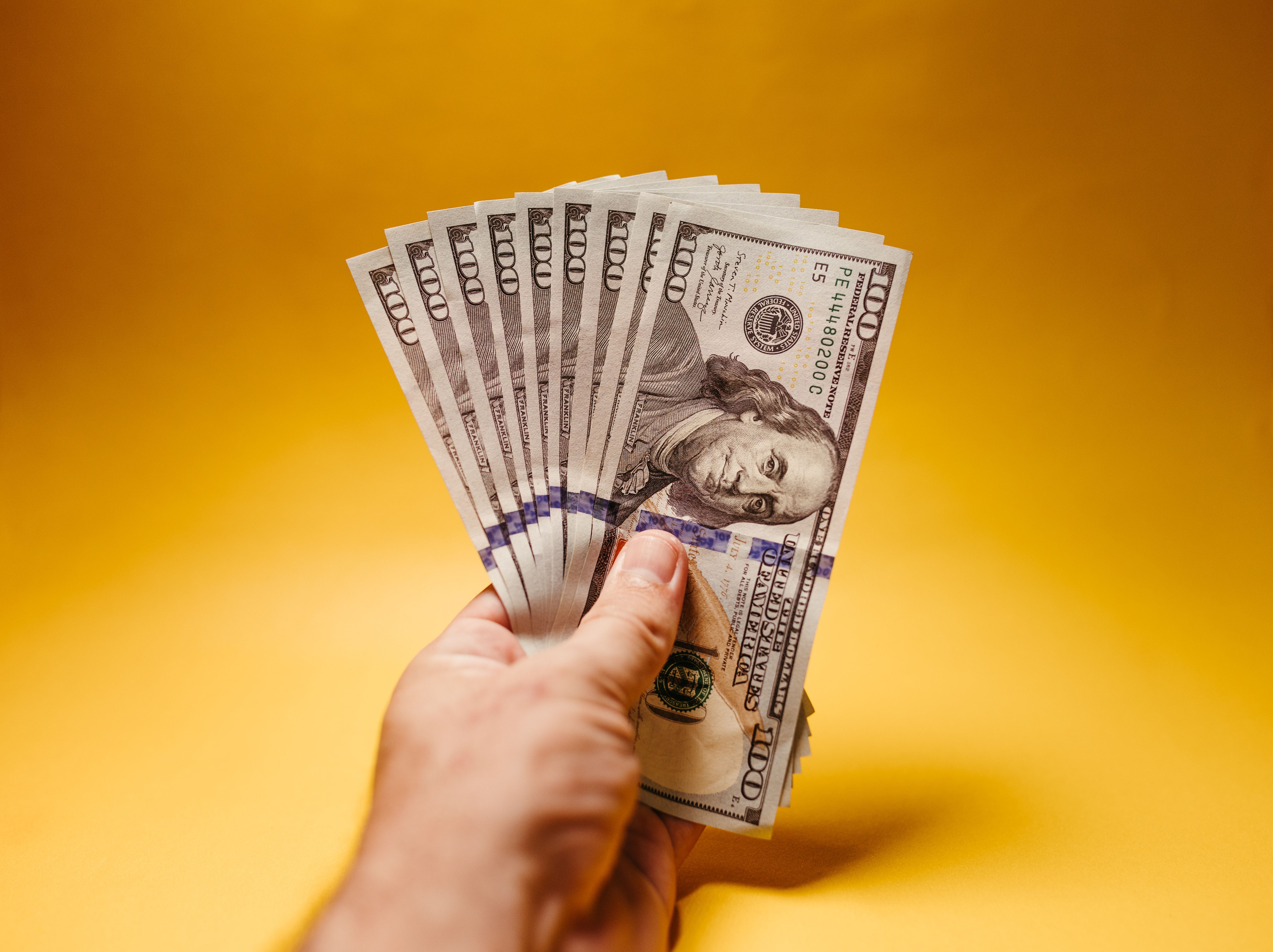A hand fans out a stack of one hundred dollar bills against a golden background.