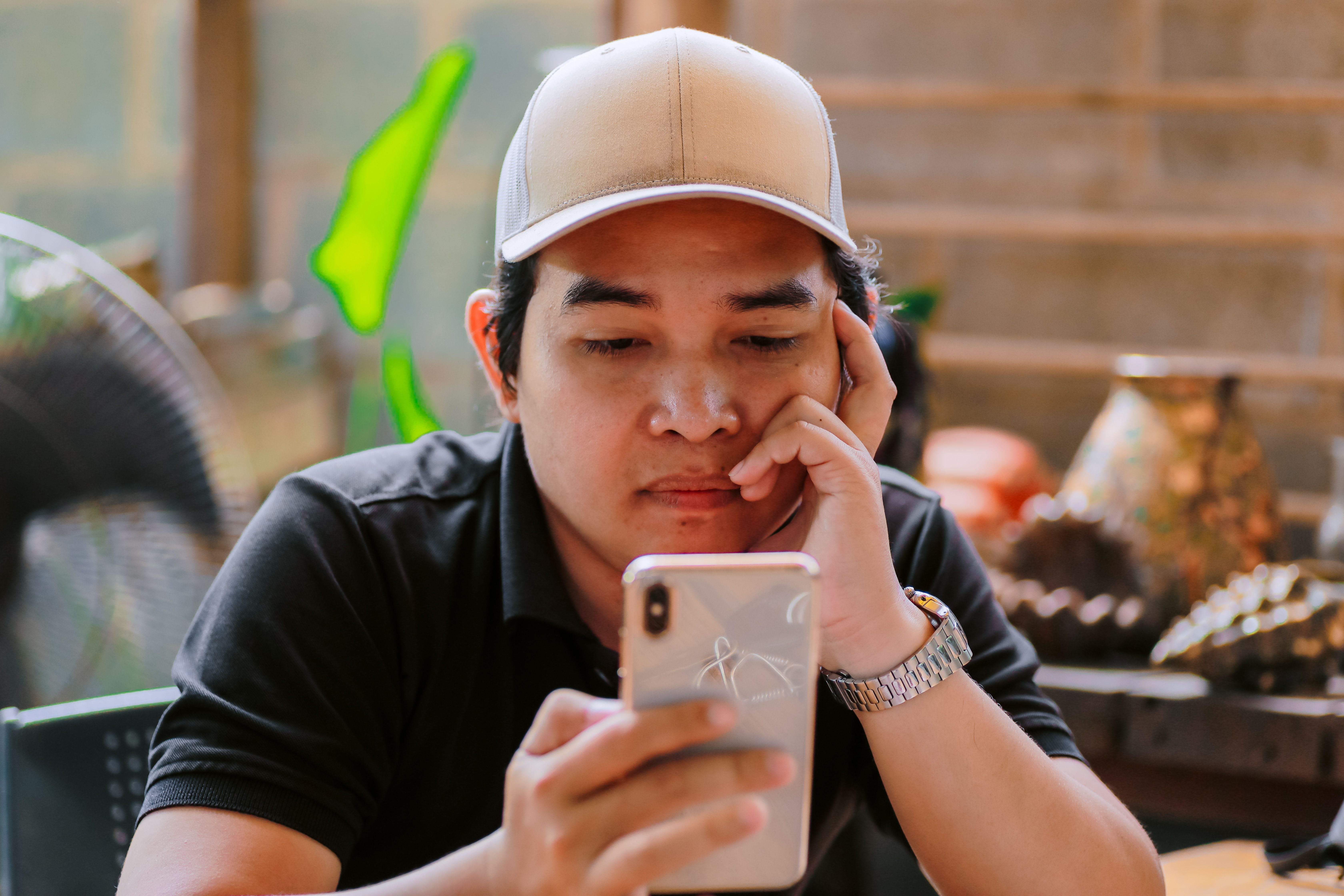 A man in a cap looks intently at his phone while resting his chin on his hand.