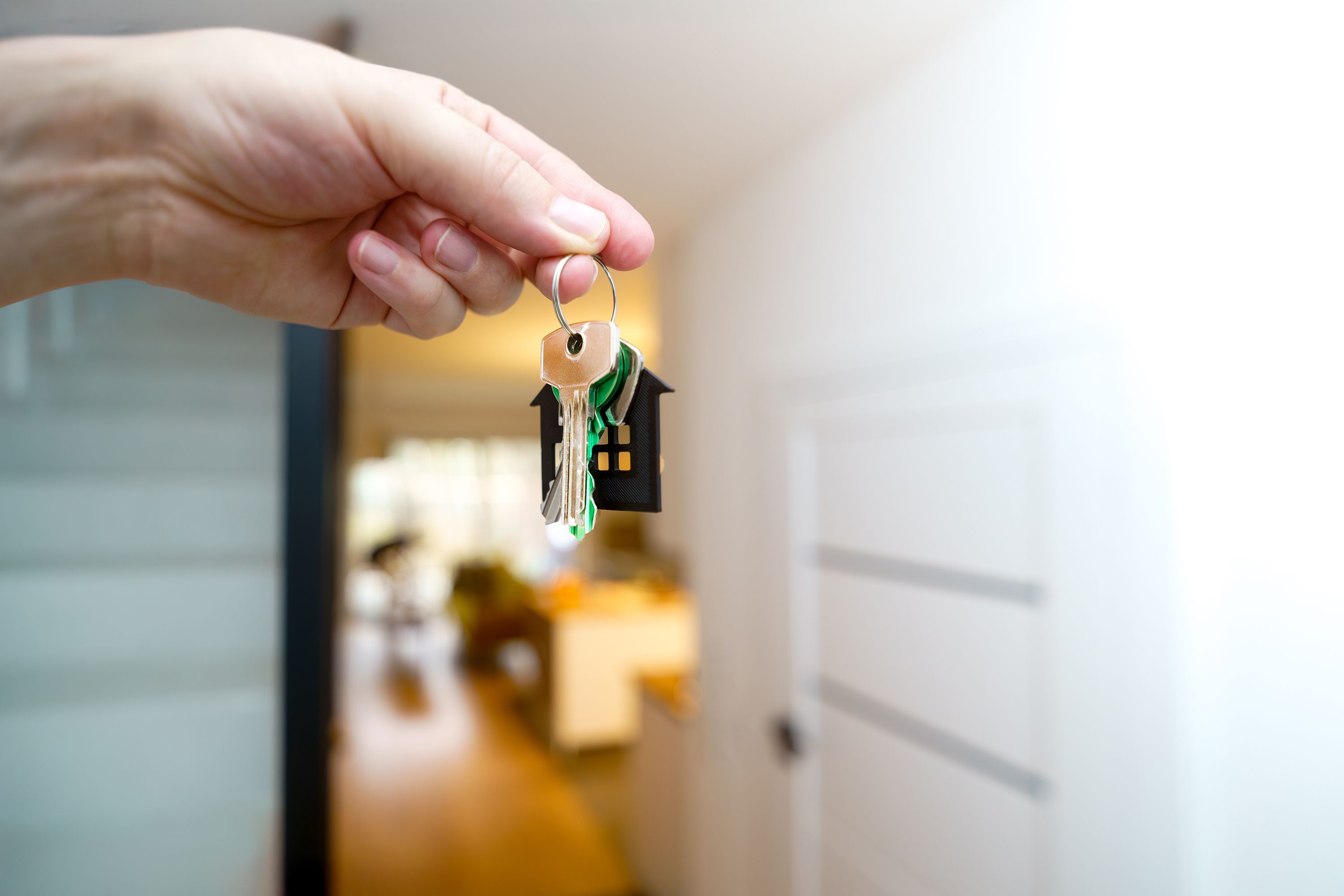 A hand holds up a set of house keys with a house-shaped keychain in front of an open front door.
