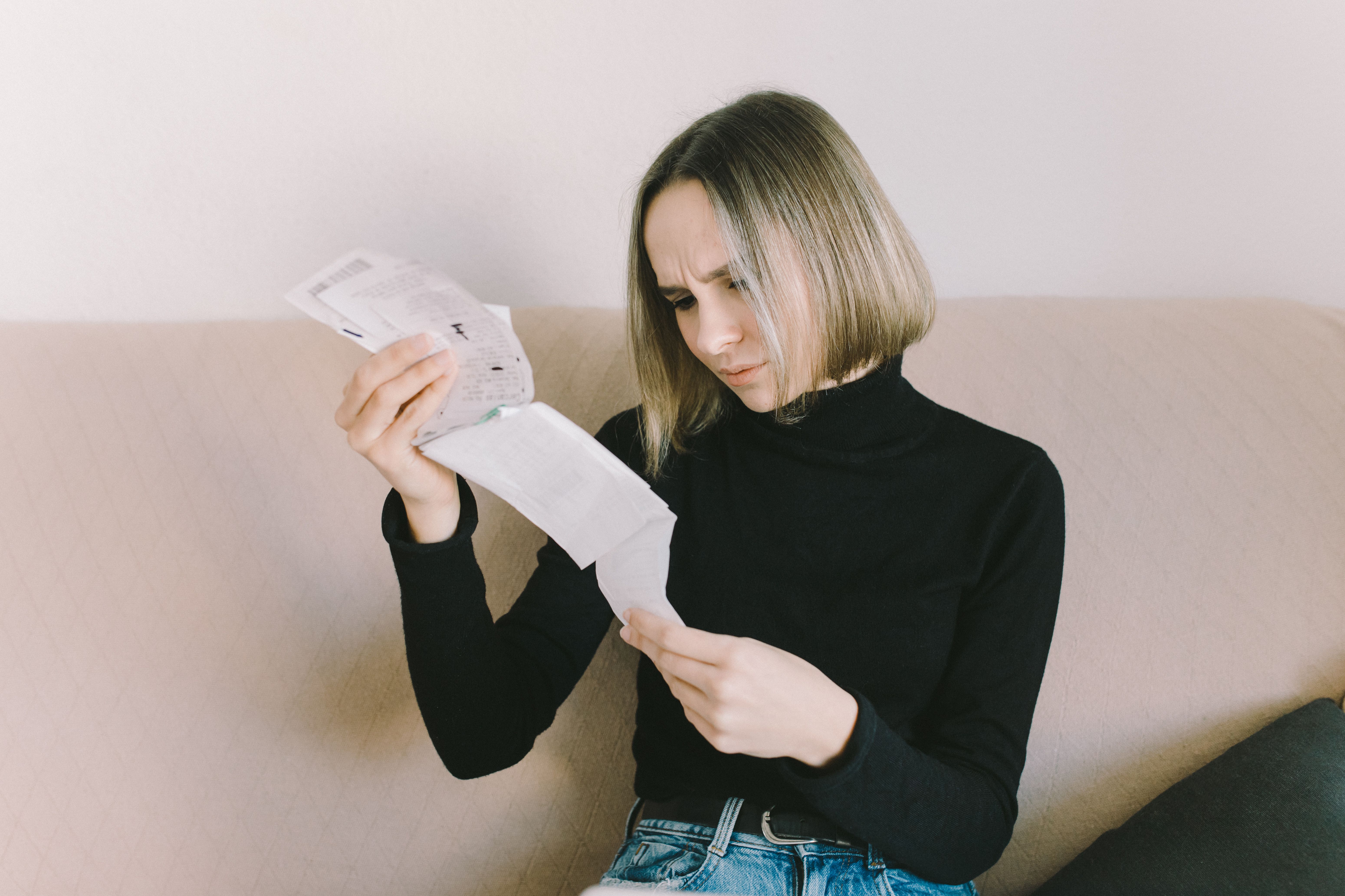 A woman sits on a couch frowning as she reads a long printed receipt or bill.