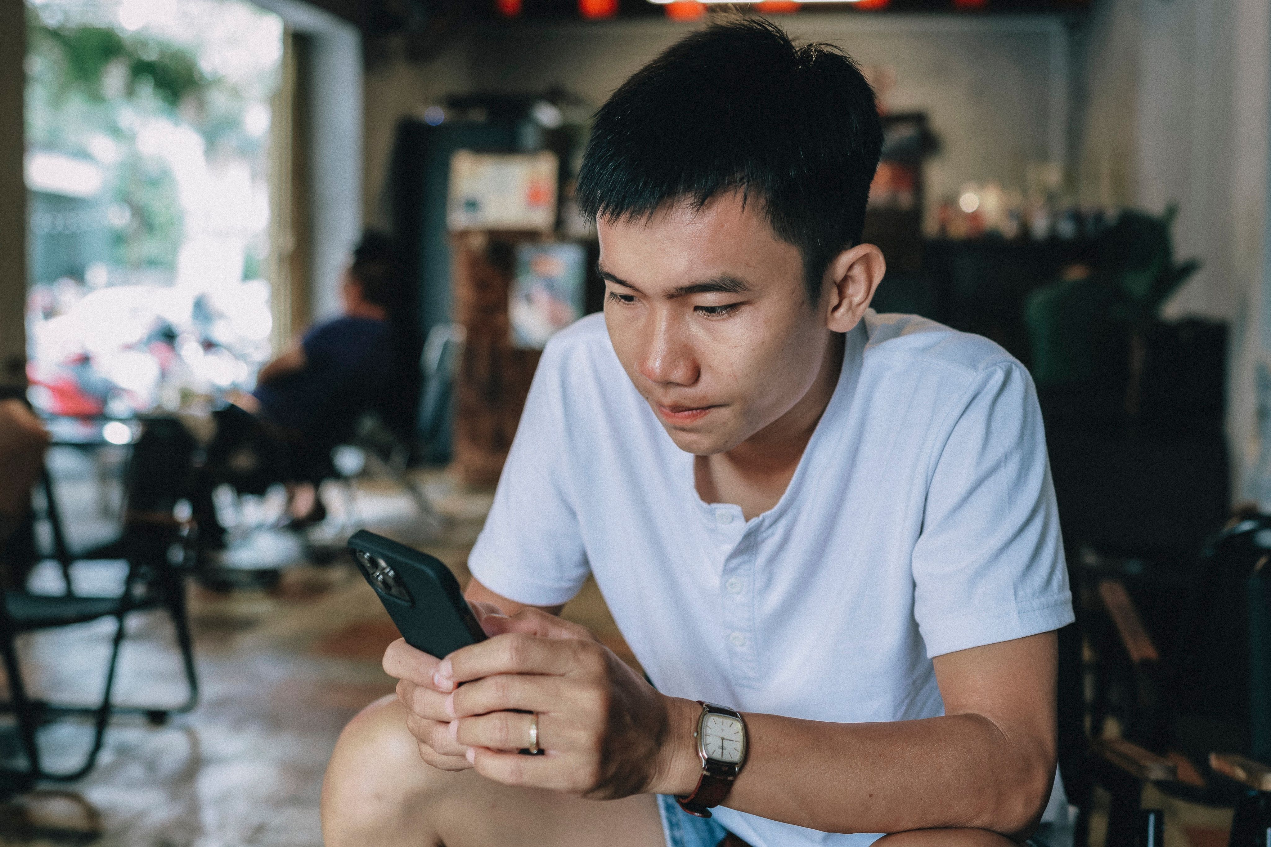 A young man sits in a café looking down at his phone with a focused expression.