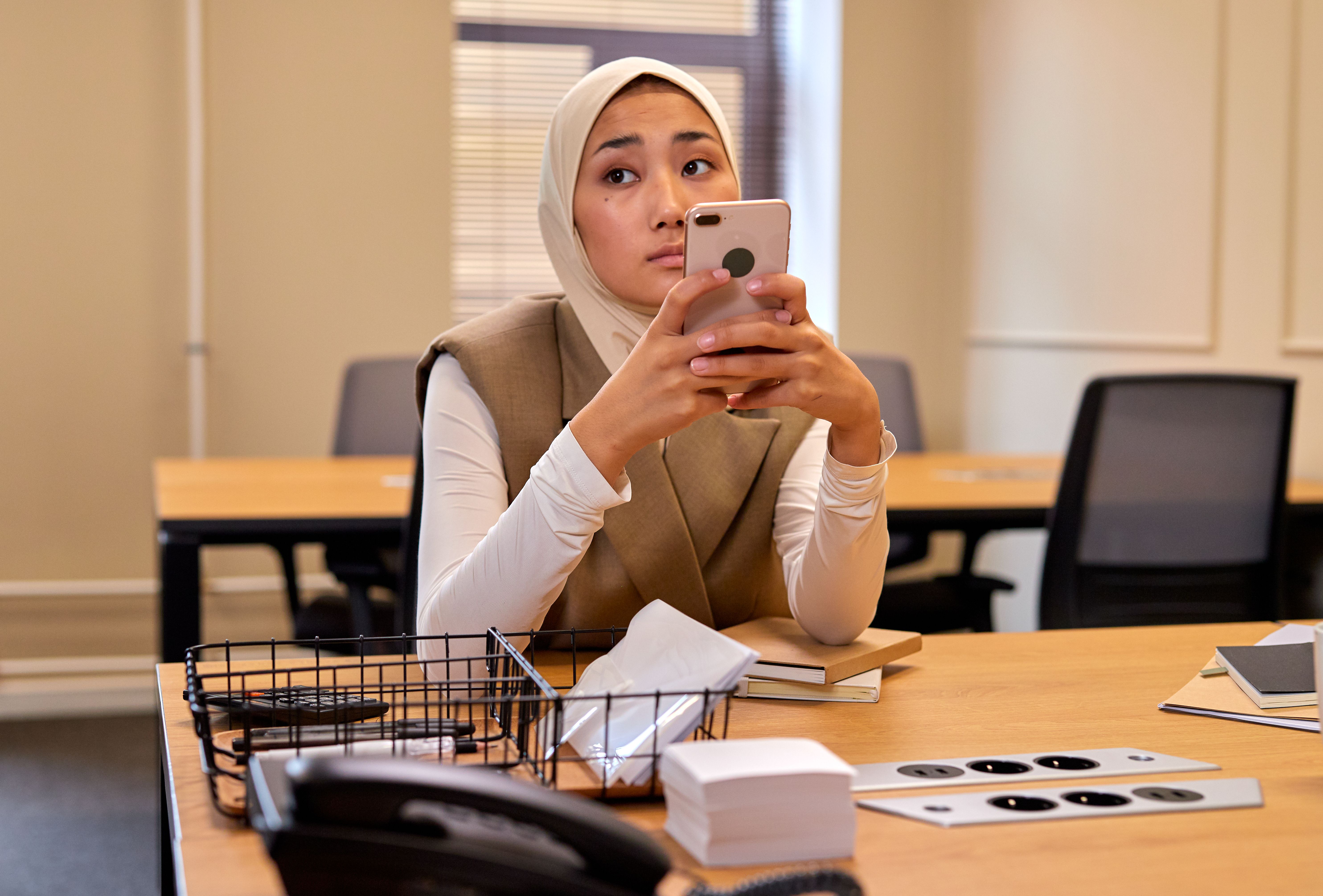 A woman in a hijab sits at an office desk, holding up her phone to take a photo.