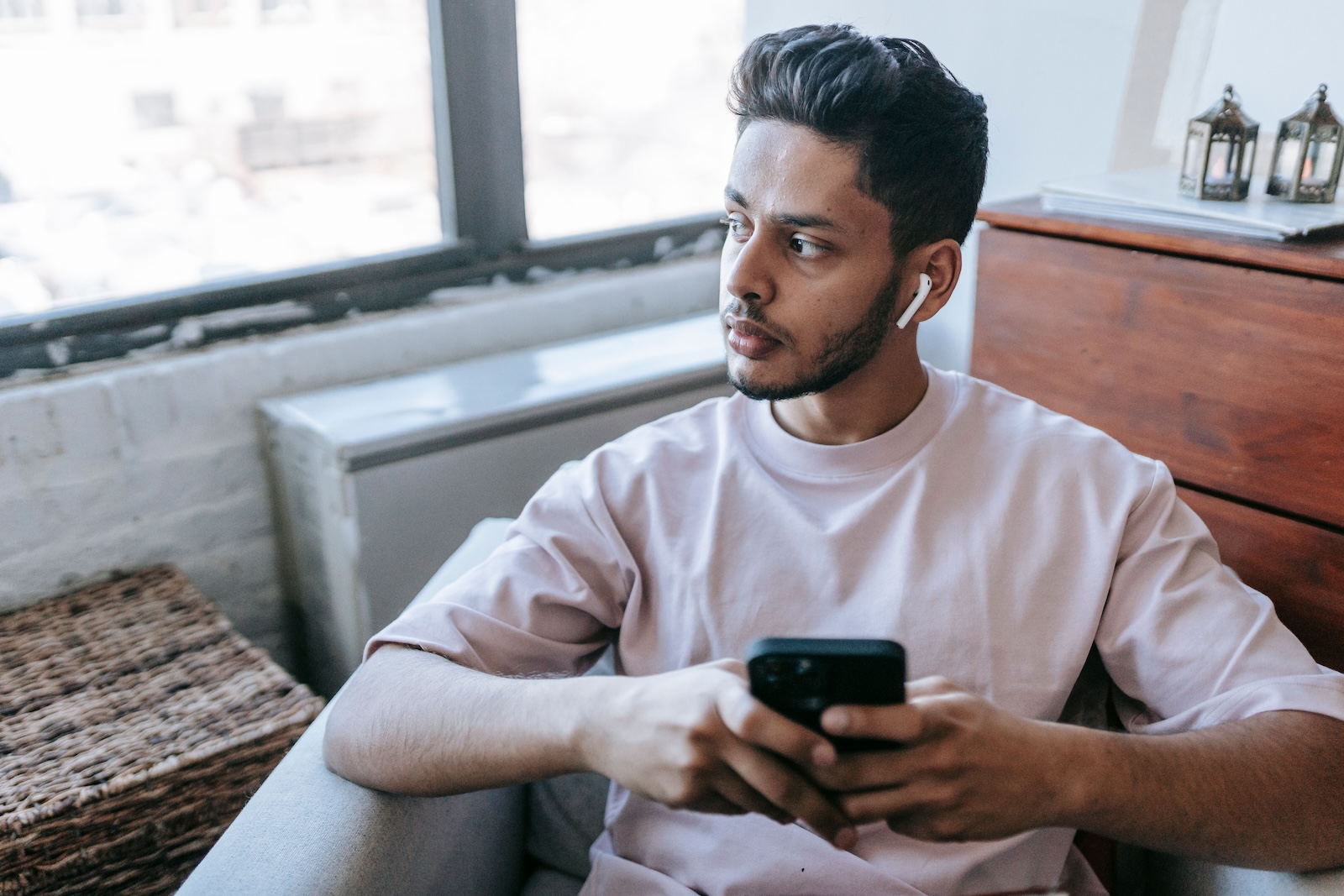 A man wearing an earbud sits by a window holding his phone, glancing off to the side.