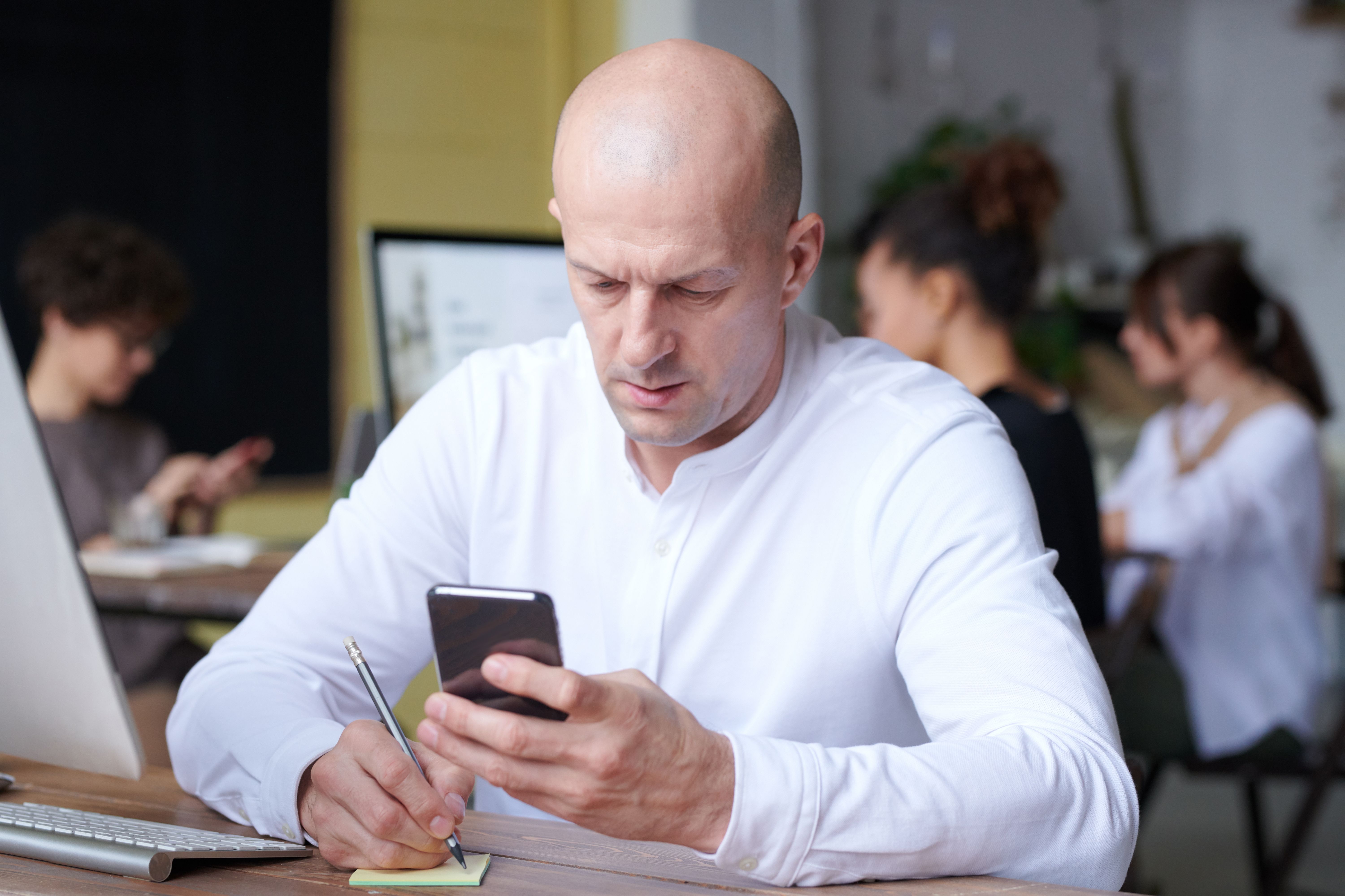 A man at a desk checks his phone while jotting a note, with coworkers talking in the background.