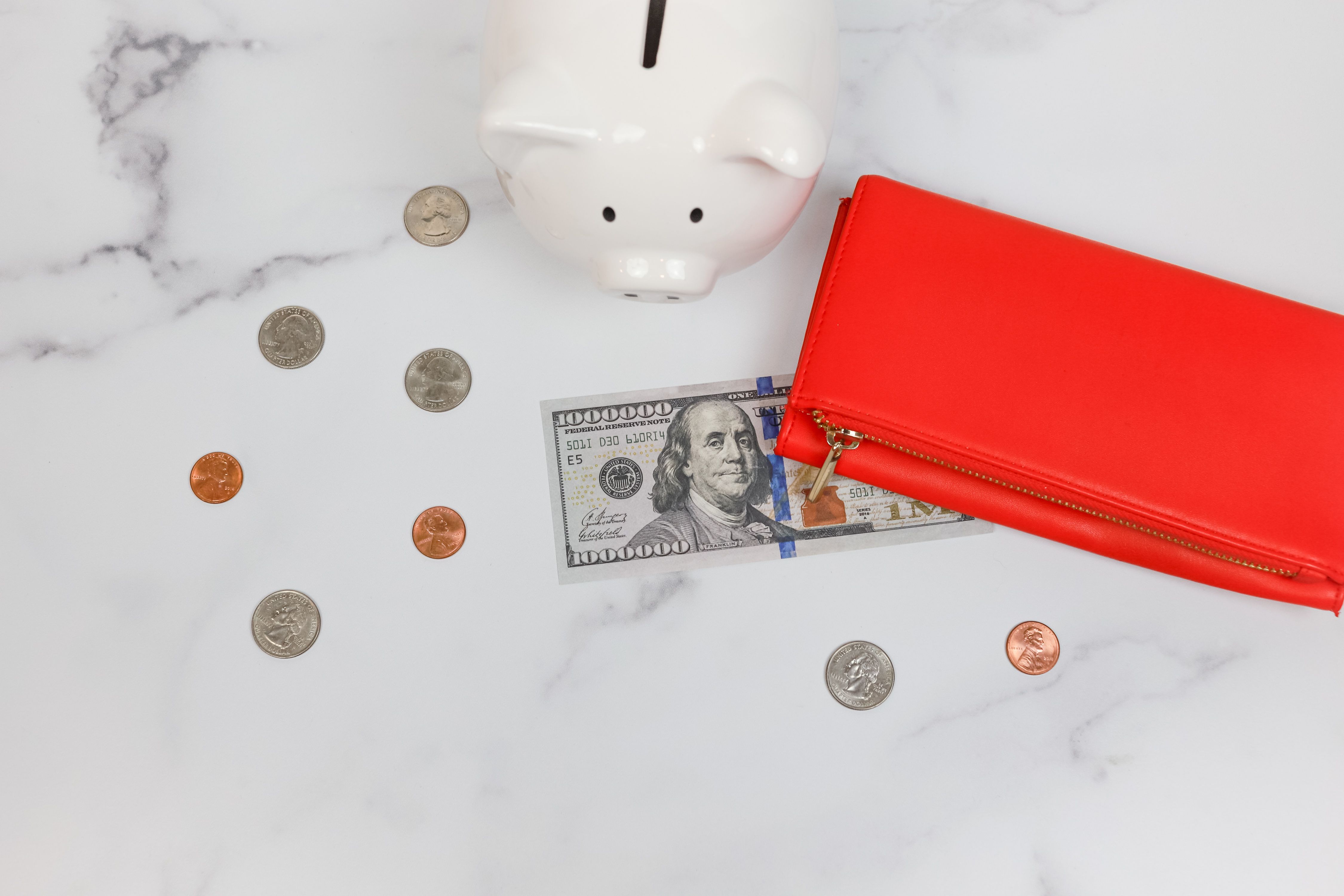 A red wallet, a hundred-dollar bill, coins, and a piggy bank arranged on a marble surface.