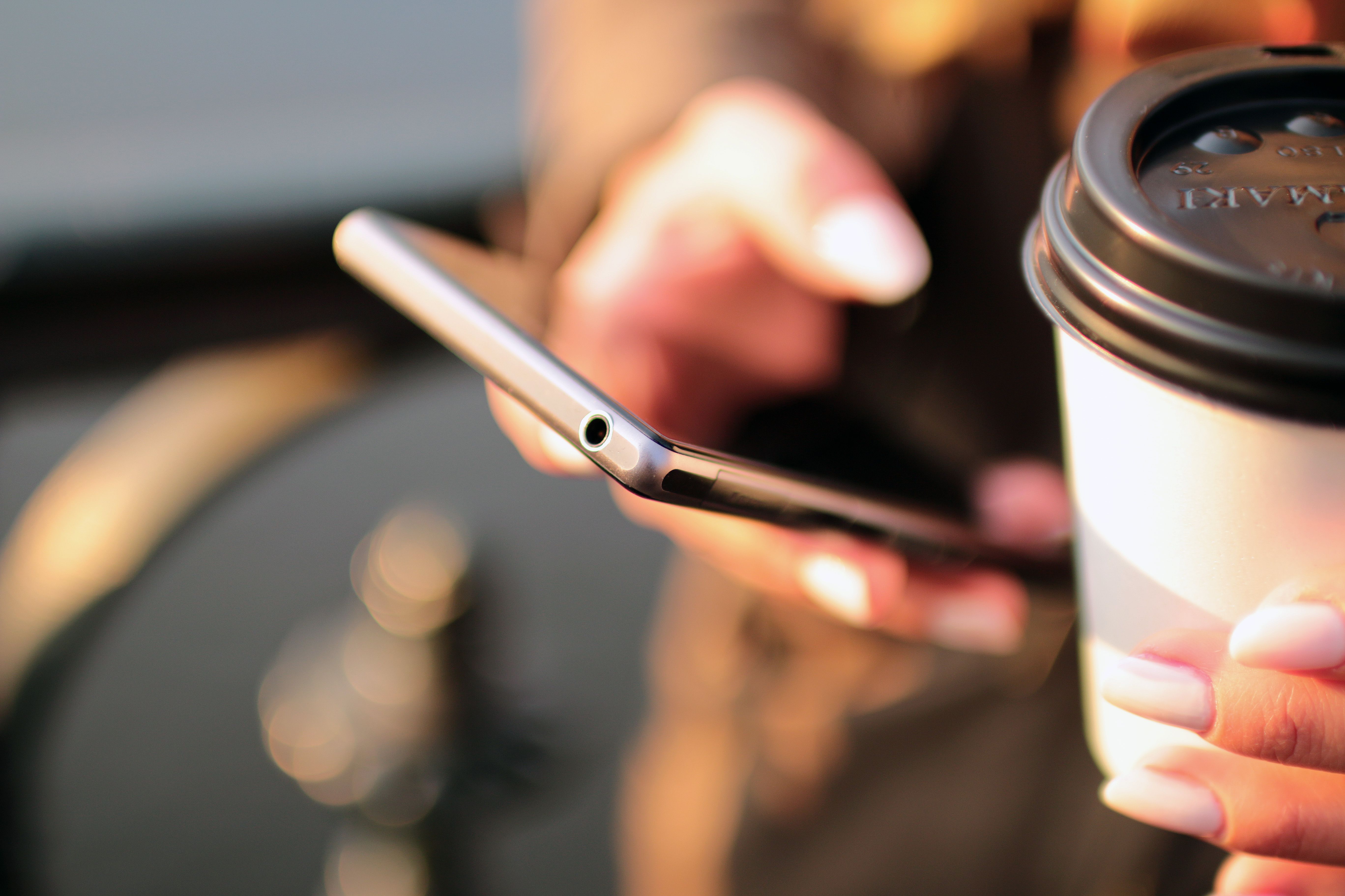 A person holding a smartphone in one hand and a to-go coffee cup with a lid in the other.