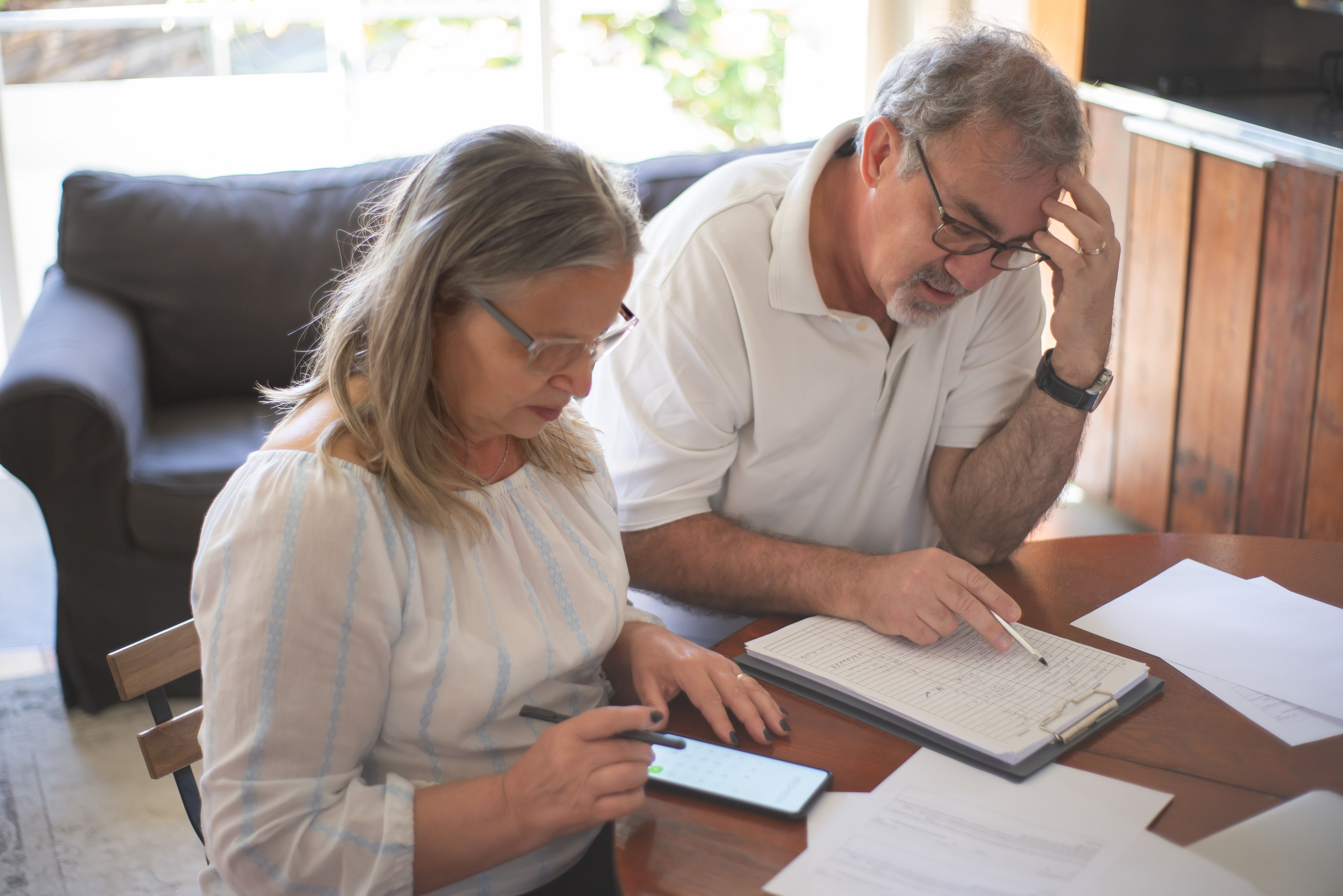 An older couple reviewing paperwork and calculating numbers on a phone at their kitchen table, looking stressed.