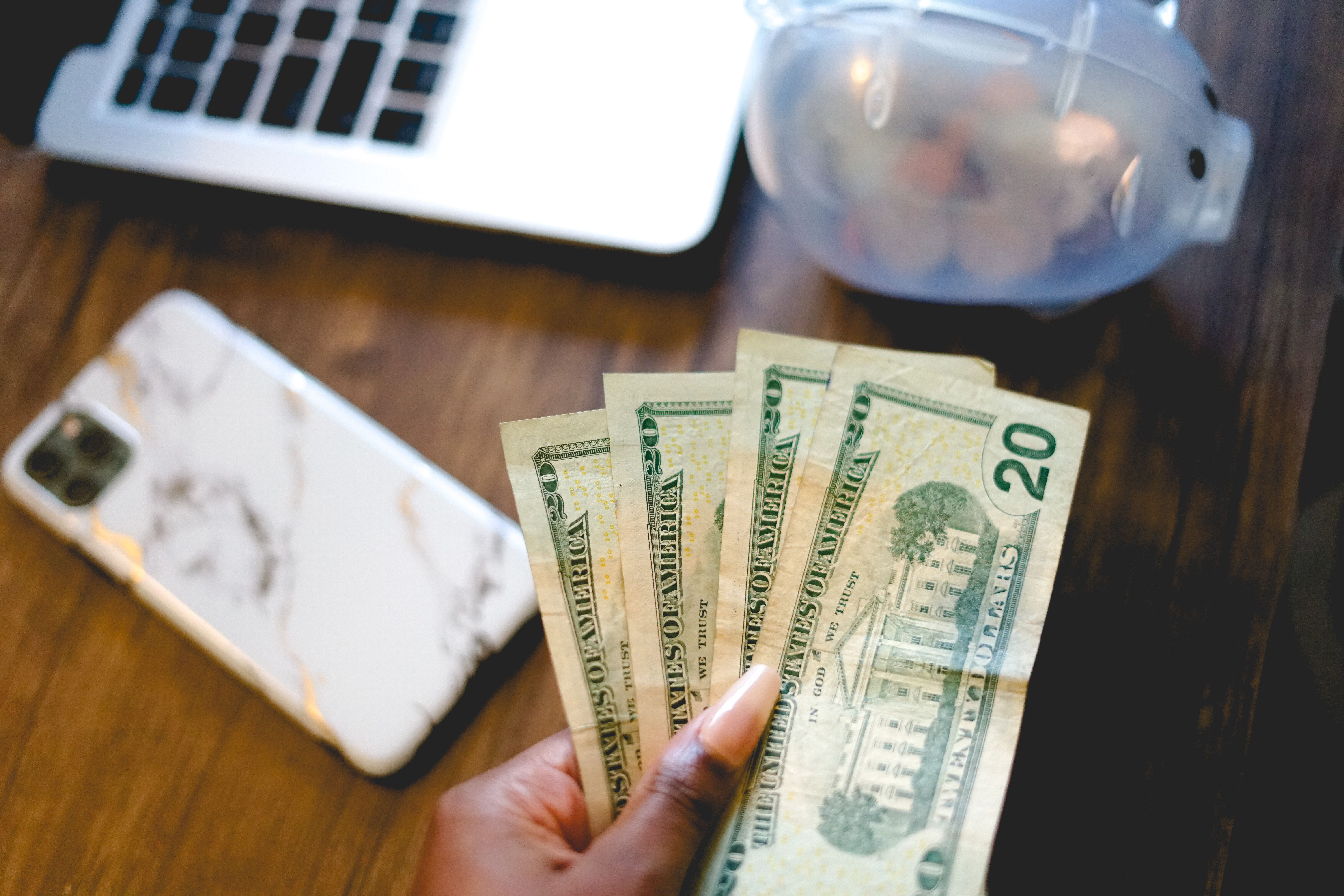 A hand fanning out several twenty-dollar bills over a desk with a phone, keyboard, and mouse.