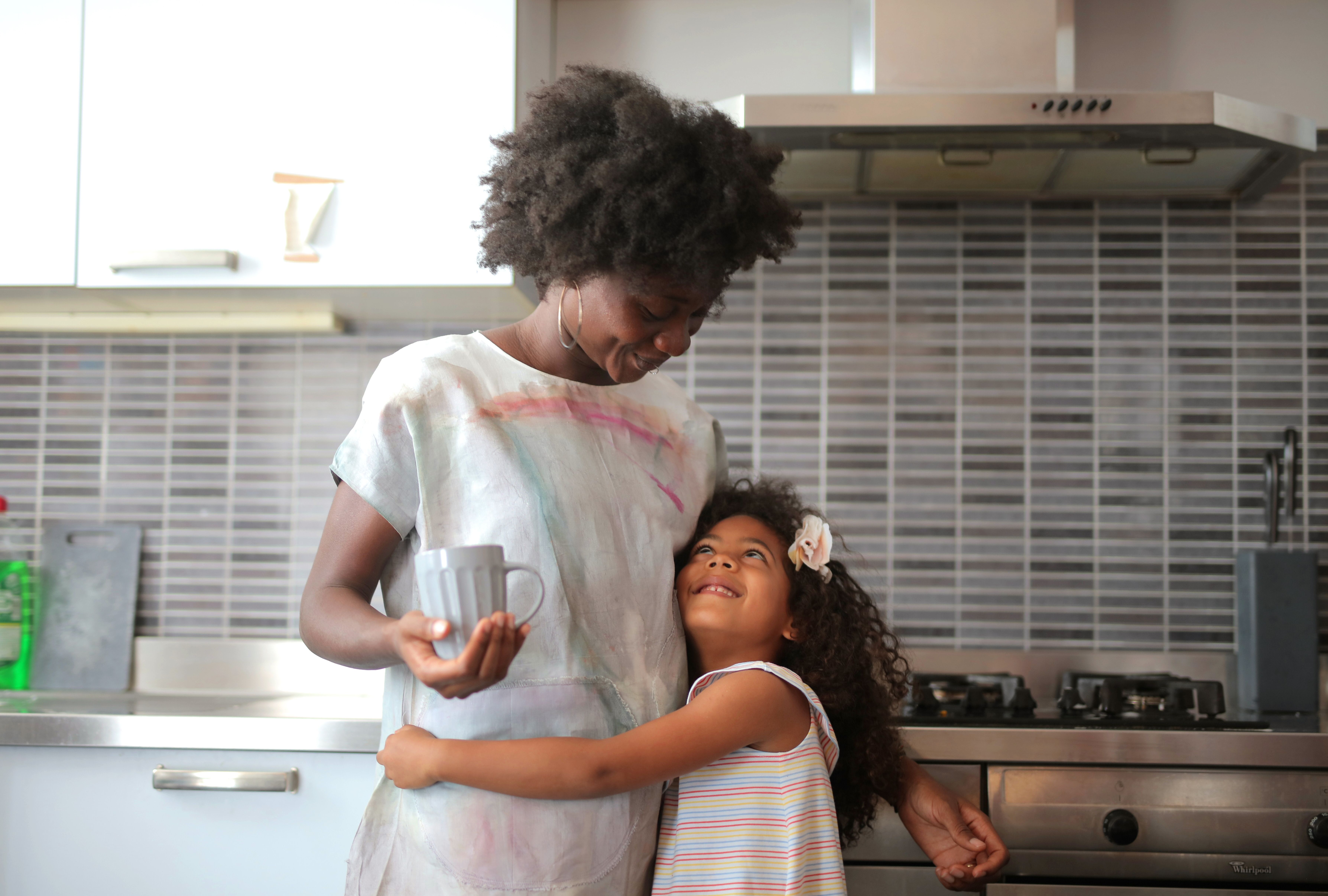 A mother holding a coffee mug while her young daughter hugs her in the kitchen, both smiling.