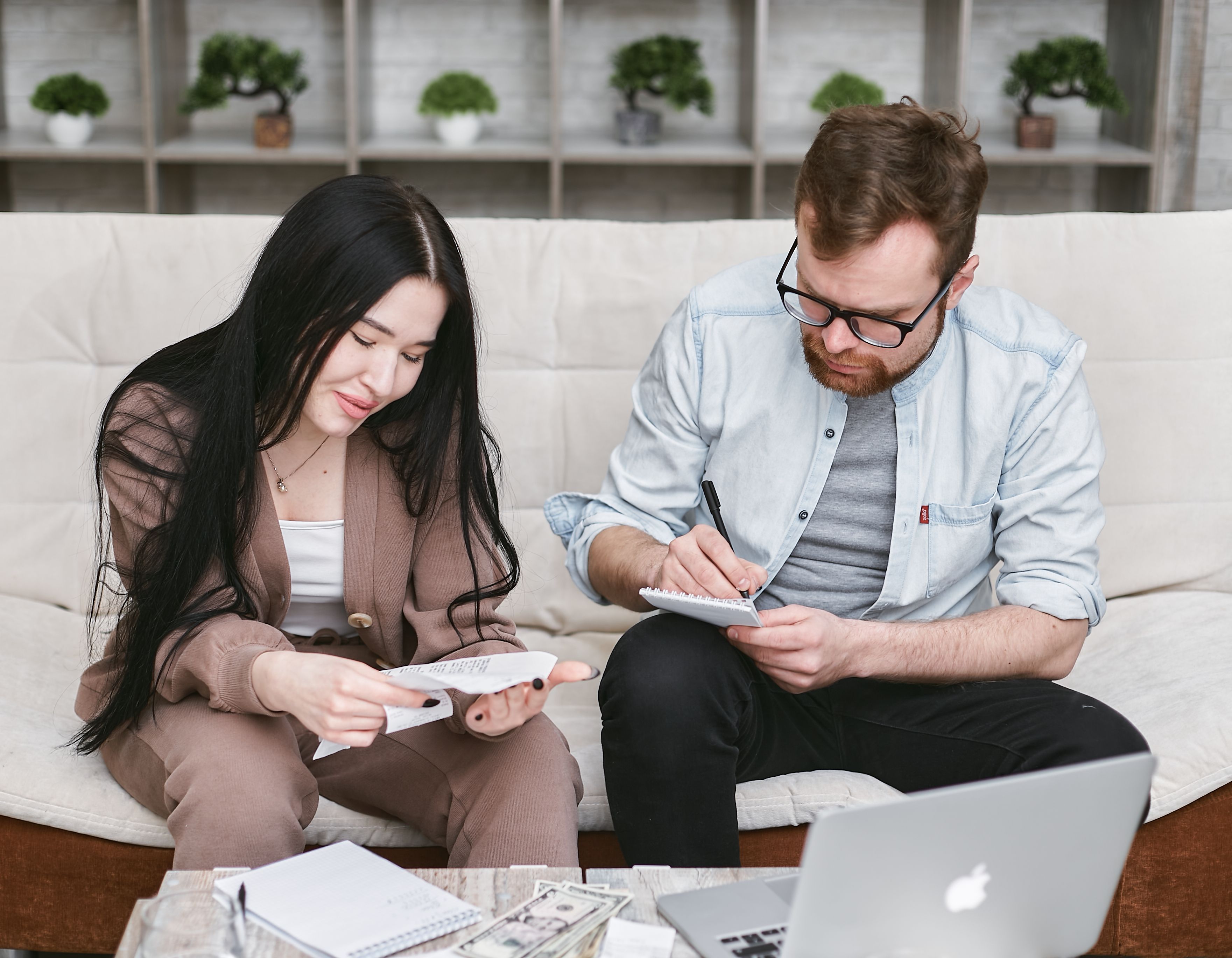 A couple sitting on a couch reviewing receipts and taking notes, with cash and a laptop on the table in front of them.
