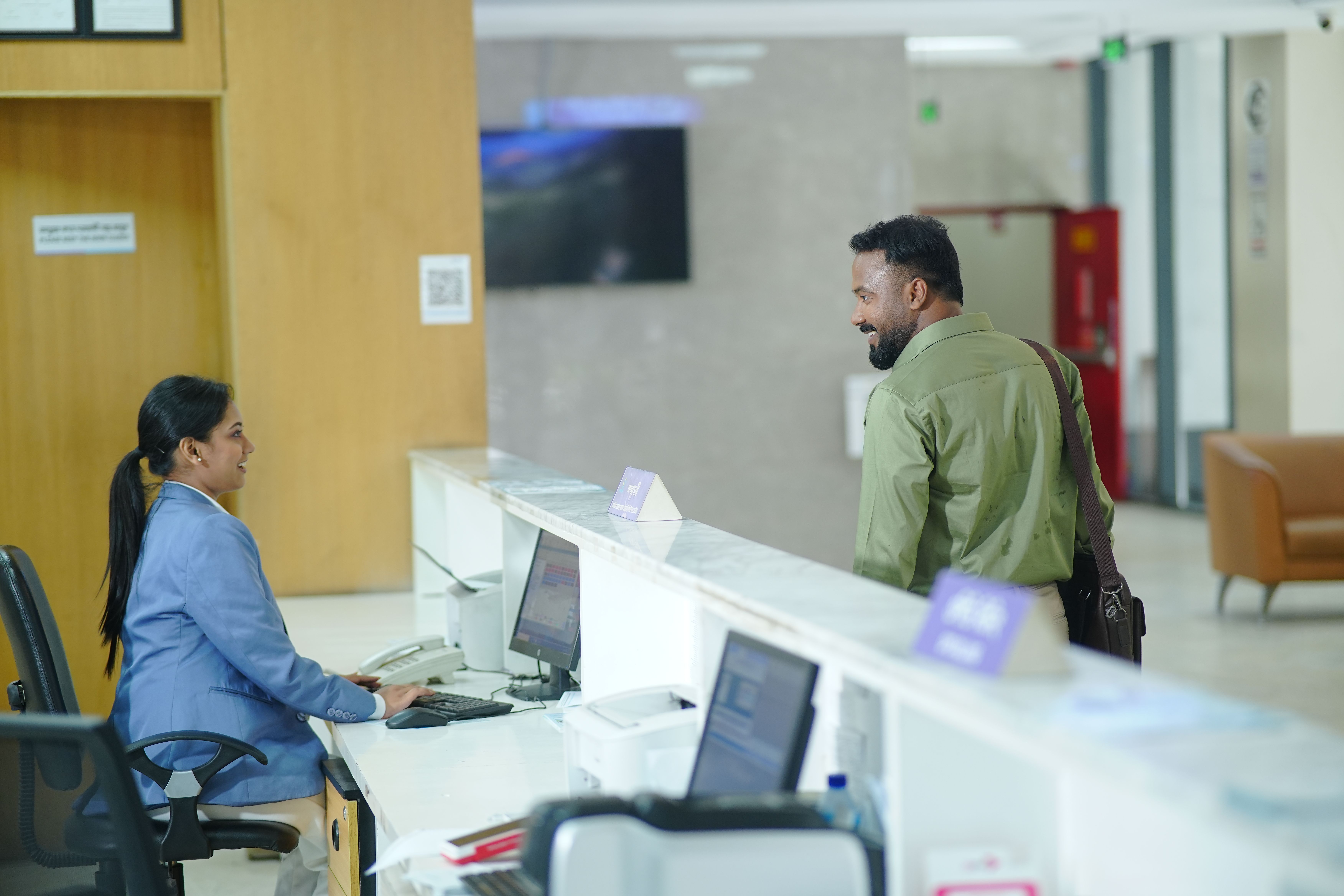 A bank teller assists a customer standing at the counter in a branch lobby.
