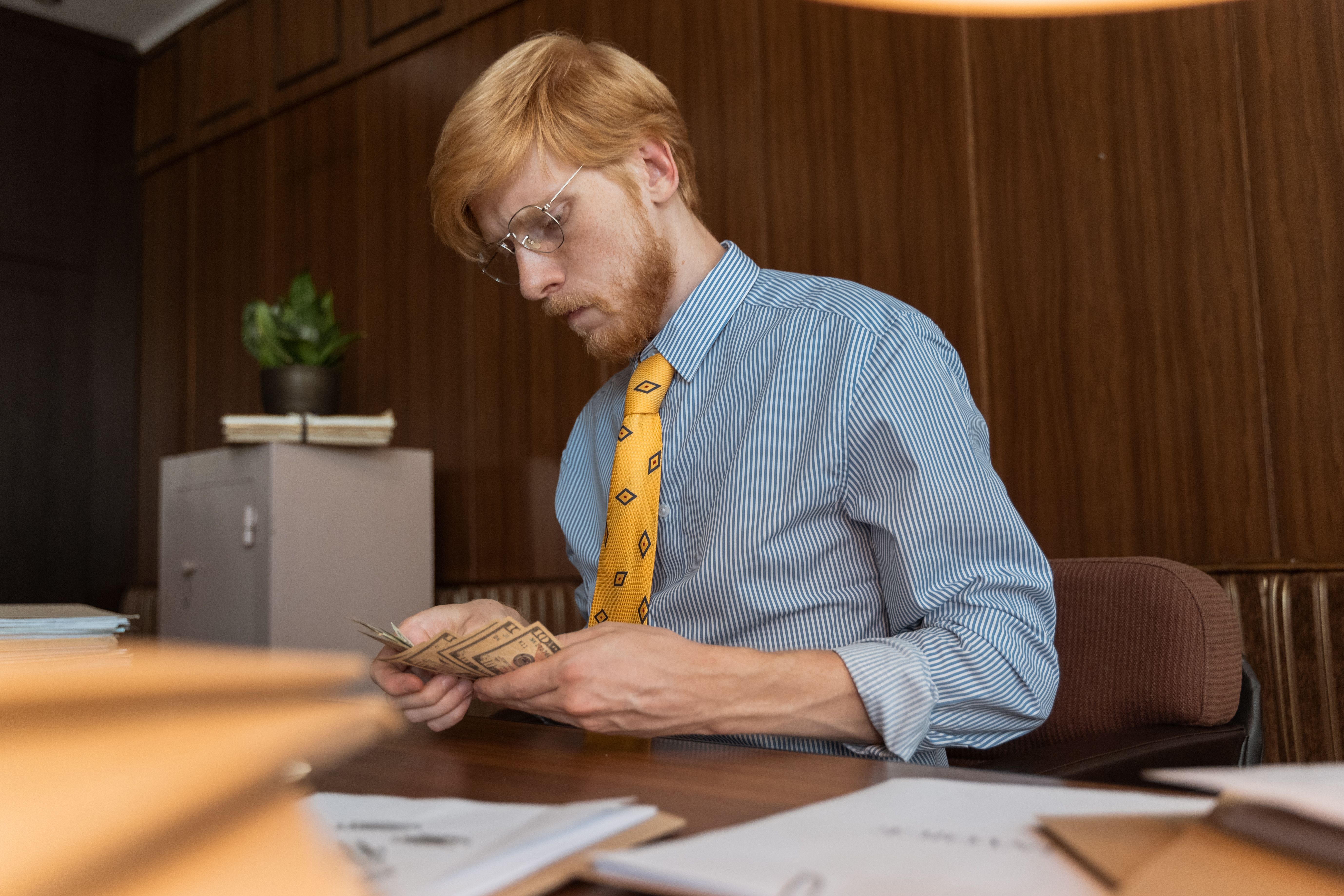 A man in a dress shirt and tie counts cash bills at a wood-paneled office desk.