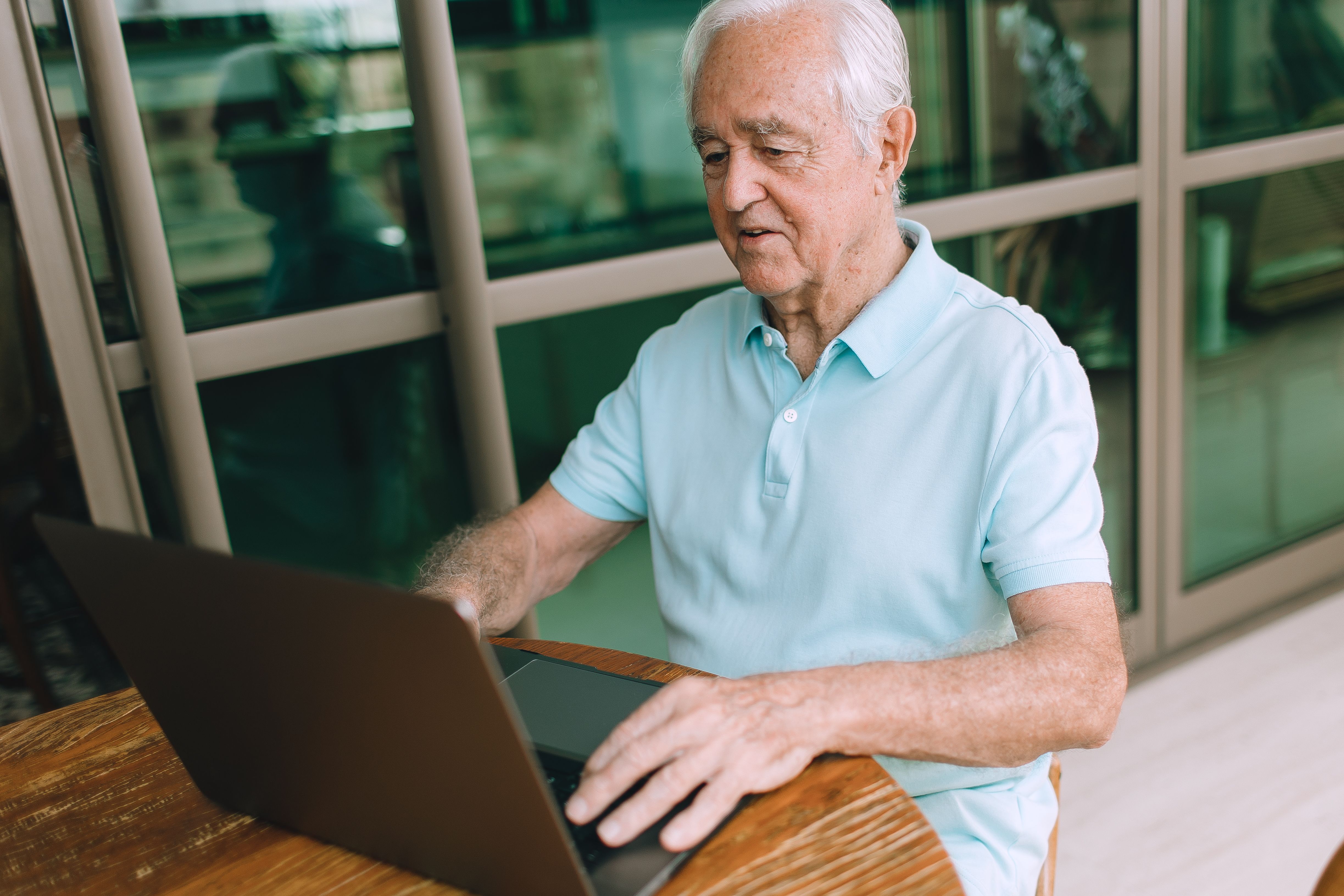 An older man types on a laptop while seated at an outdoor table.