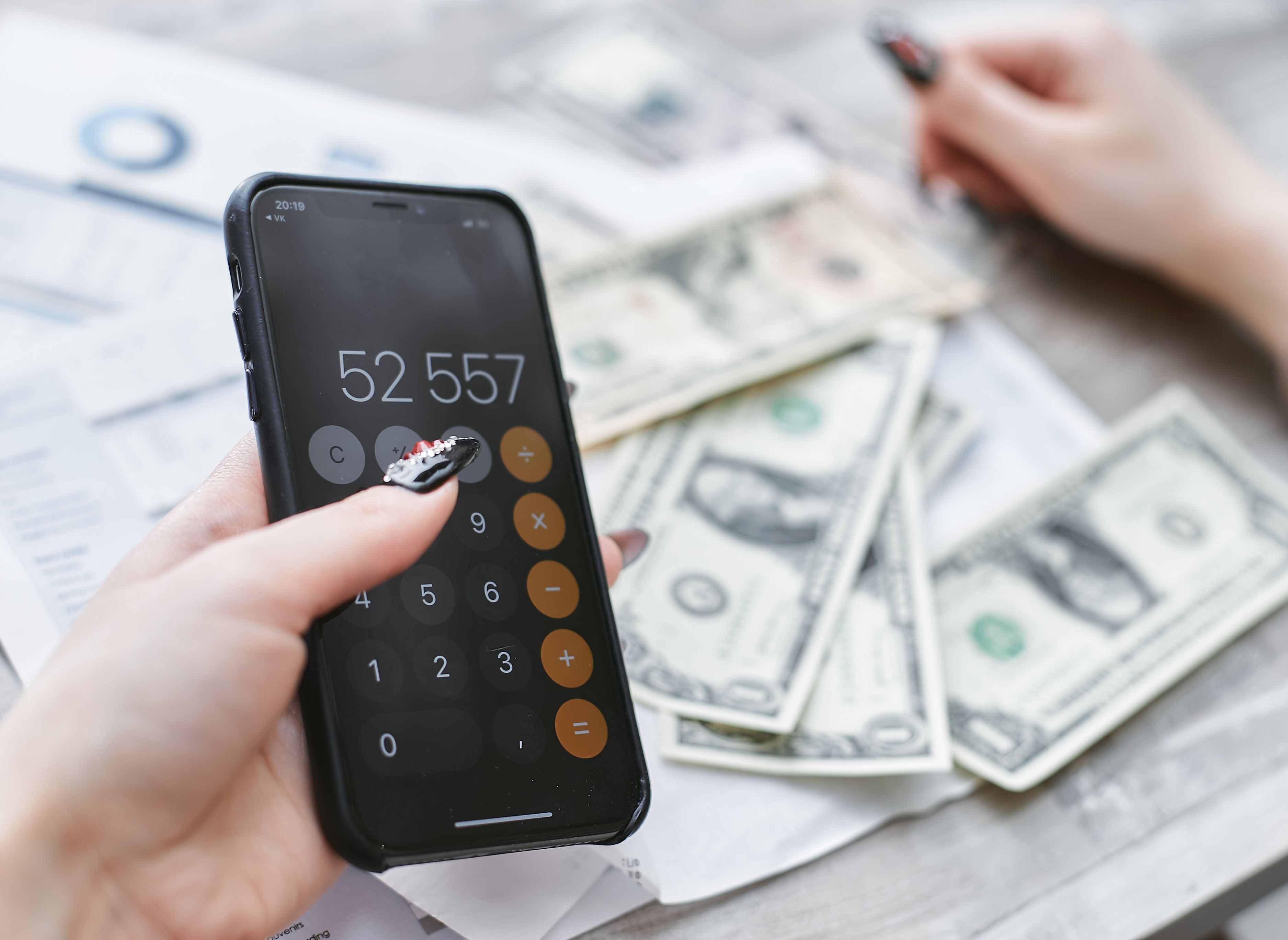 A hand uses a phone's calculator app next to cash and paperwork on a table.