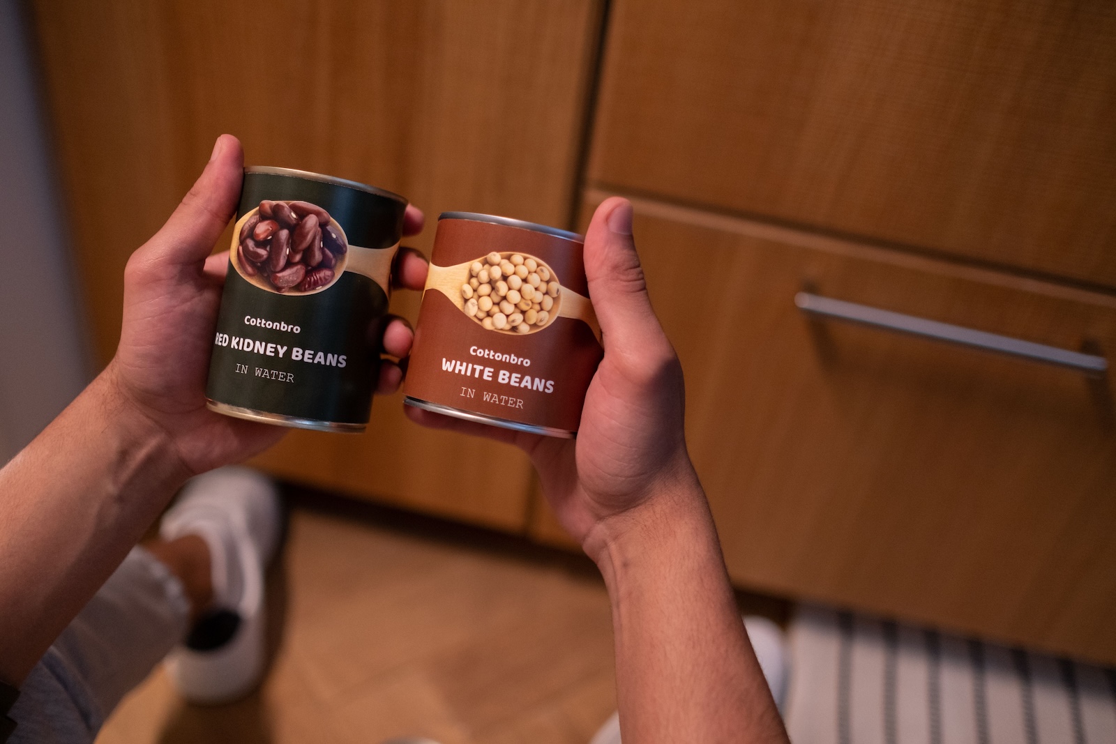 Hands hold a can of red kidney beans and a can of white beans in a kitchen.