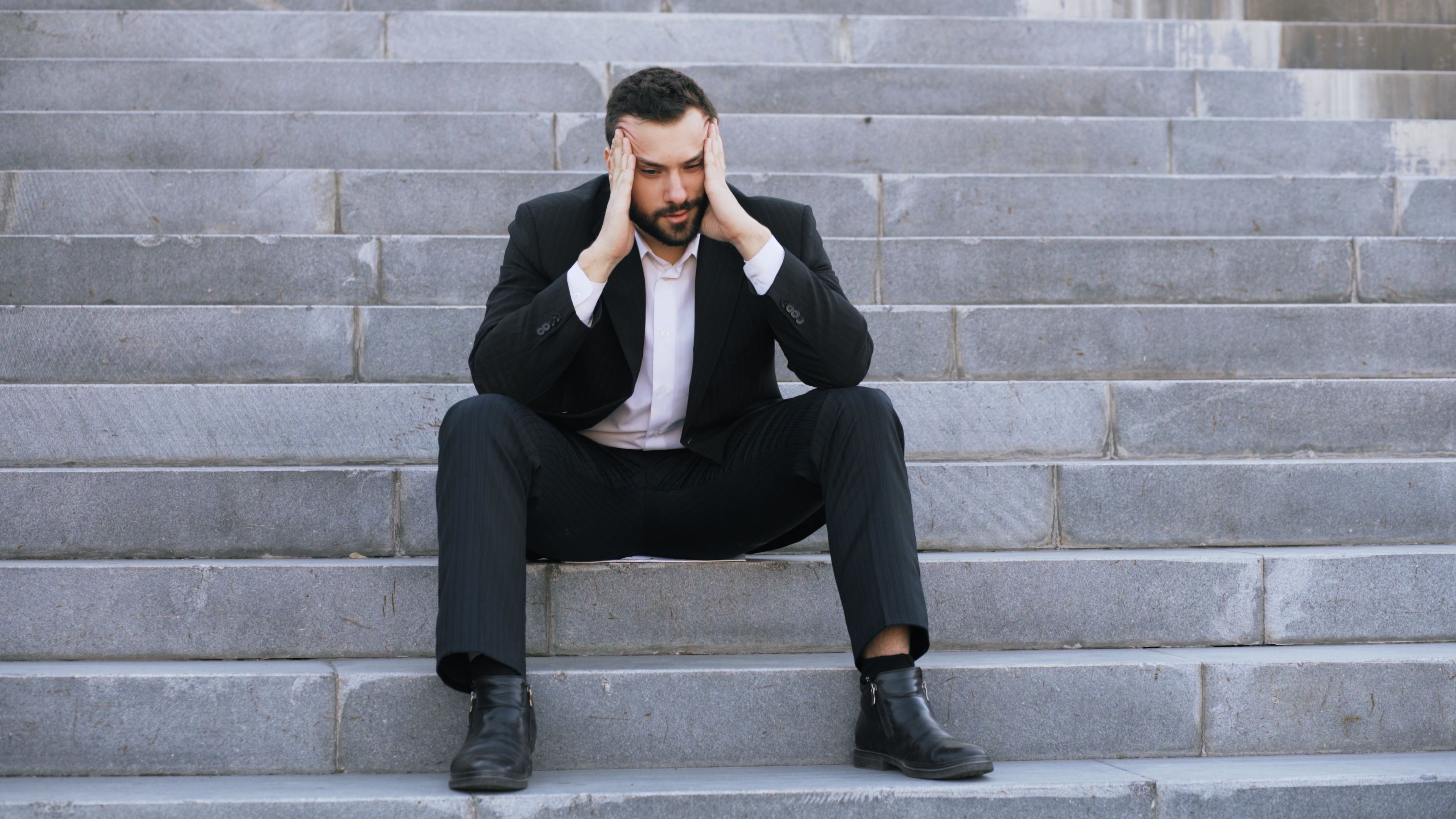 Photo of a man in a dark suit sitting on outdoor stone steps with his head in his hands.