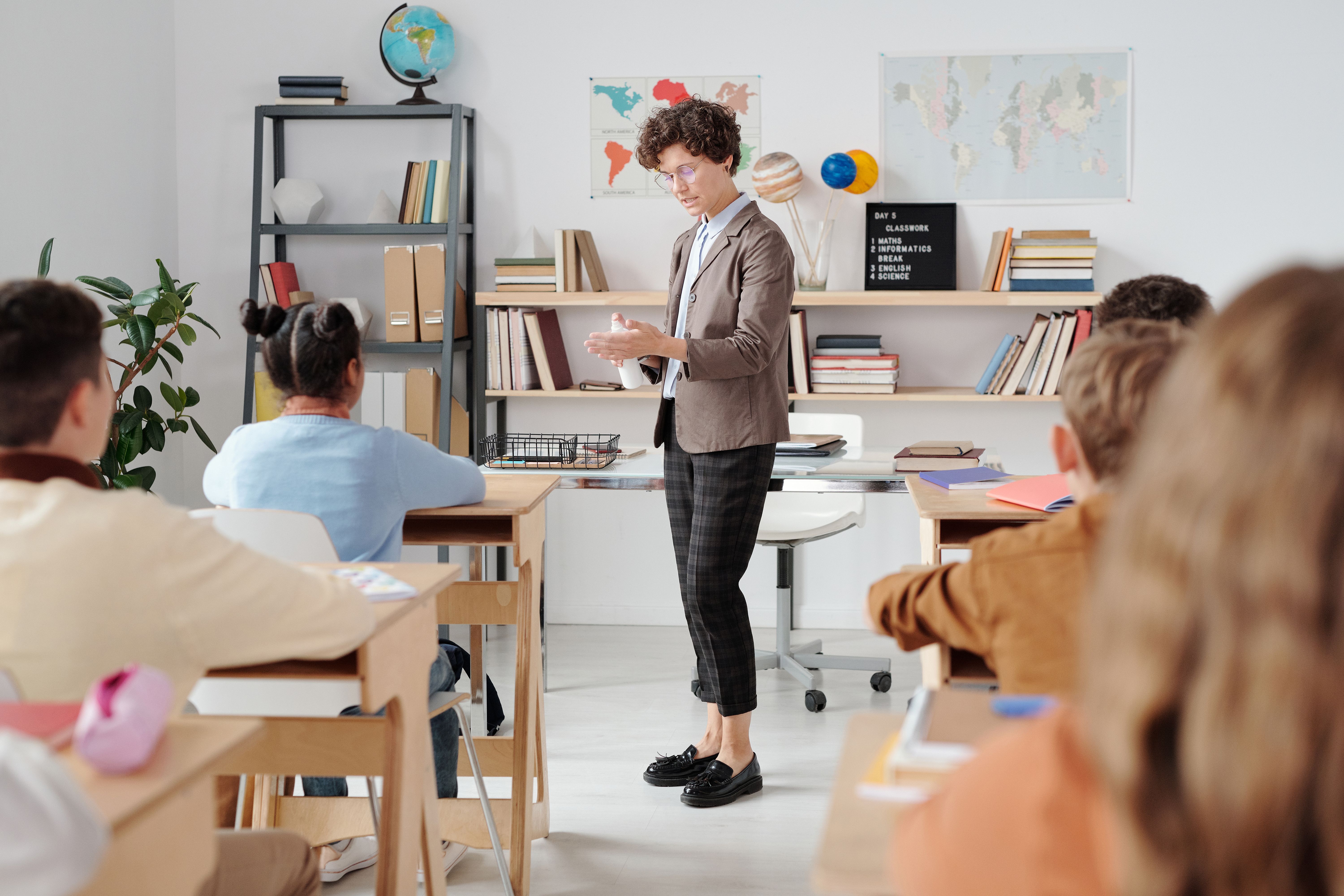 Photo of a teacher using hand sanitizer at the front of a classroom while students sit at their desks.