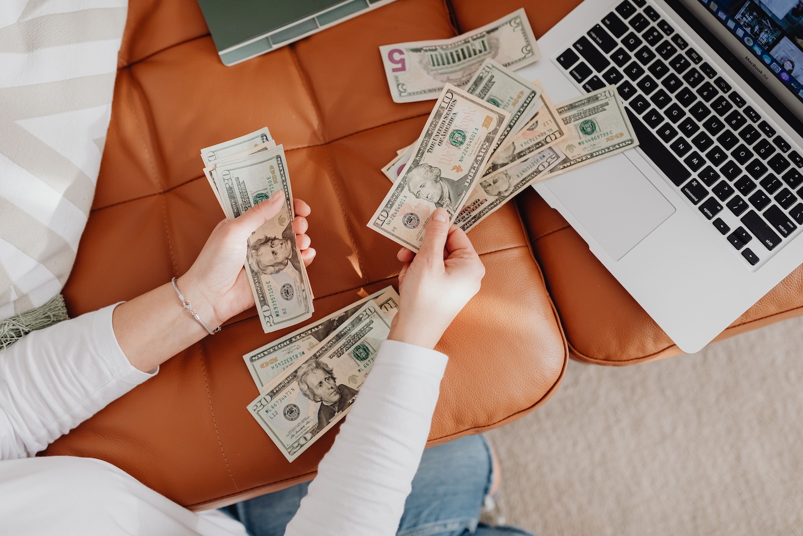 Photo of a person counting cash on an orange leather chair beside an open laptop.