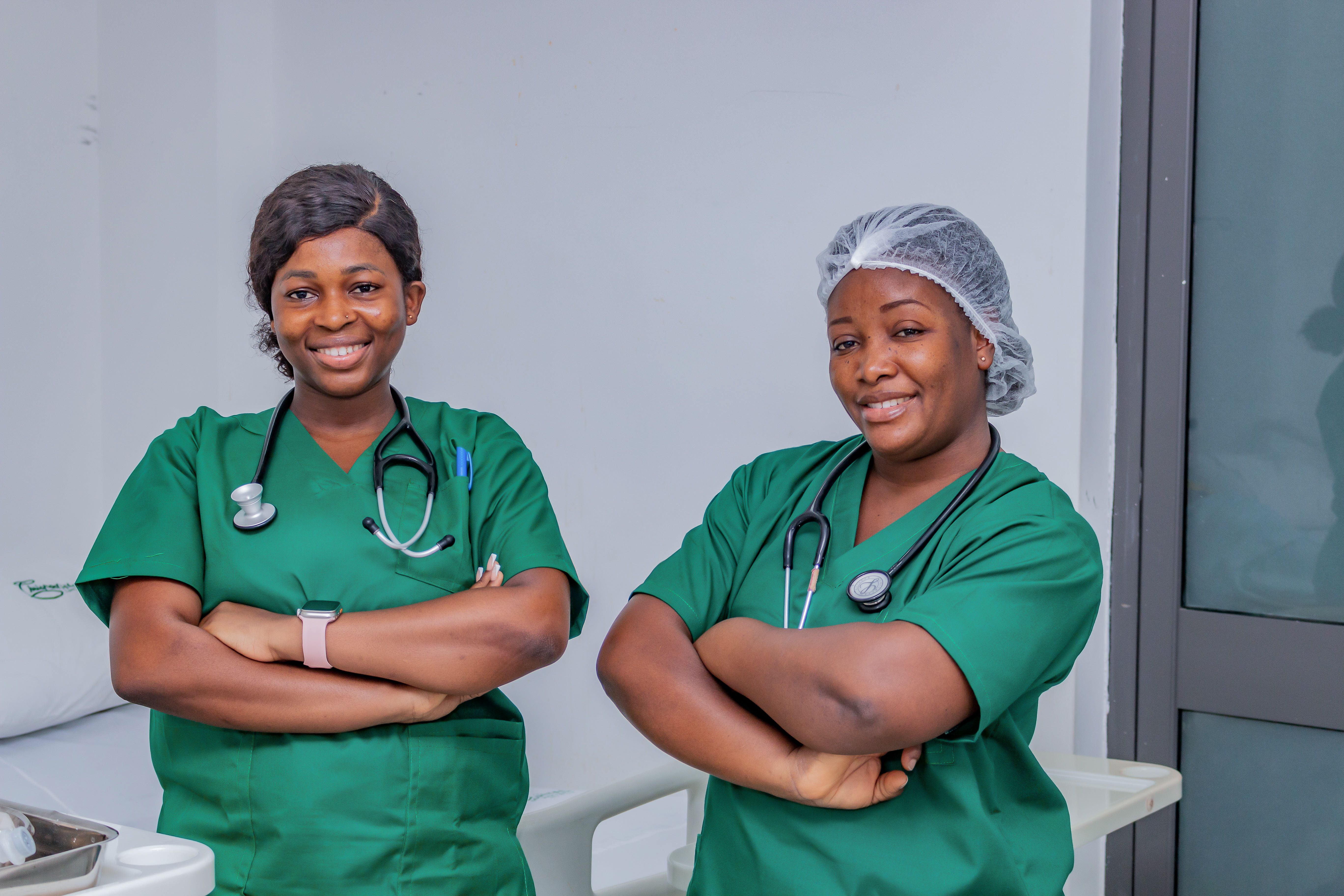 Photo of two smiling nurses in green scrubs with their arms crossed in a hospital room.