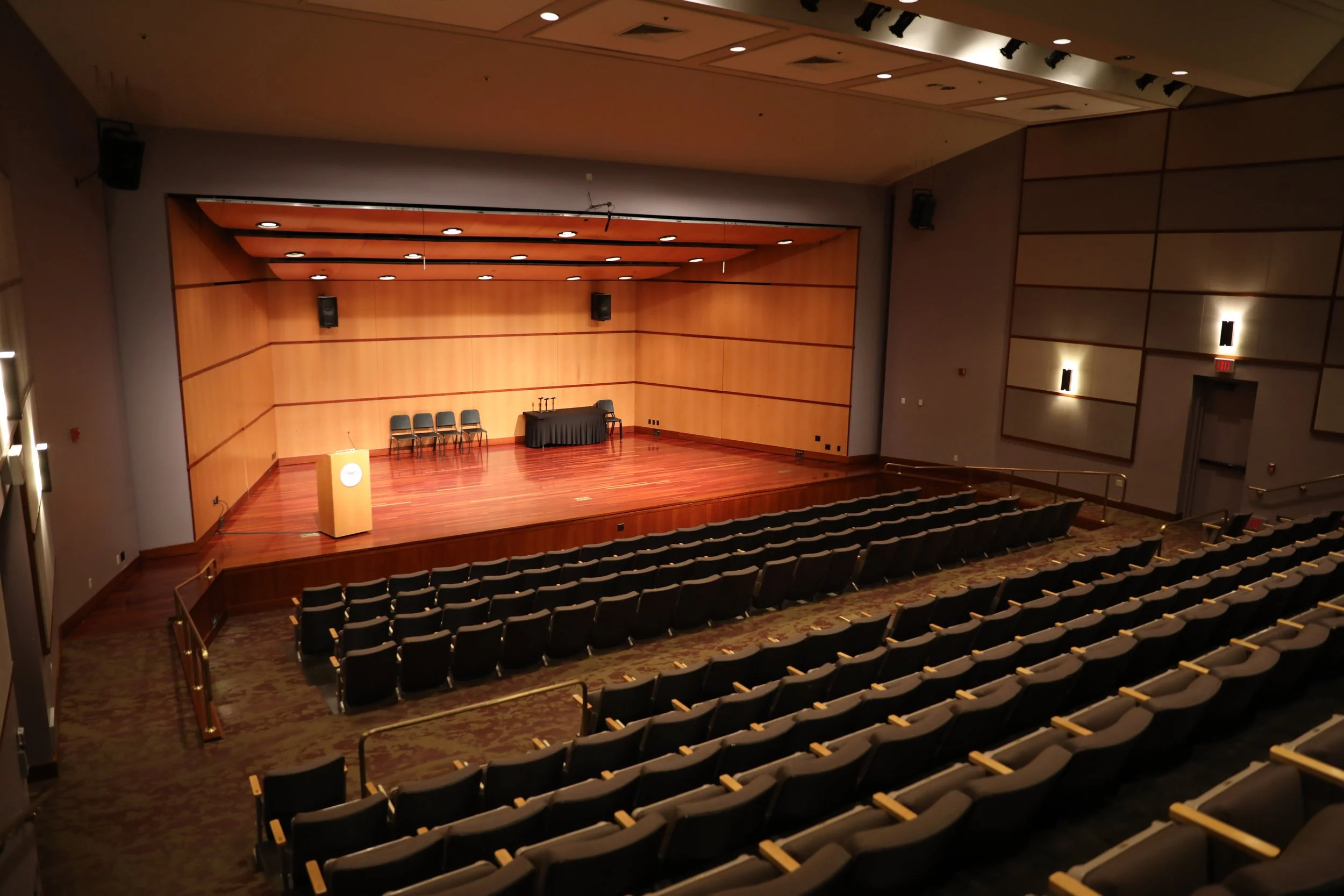 Empty auditorium with tiered seating facing a stage with a desk, microphone, and chairs.
