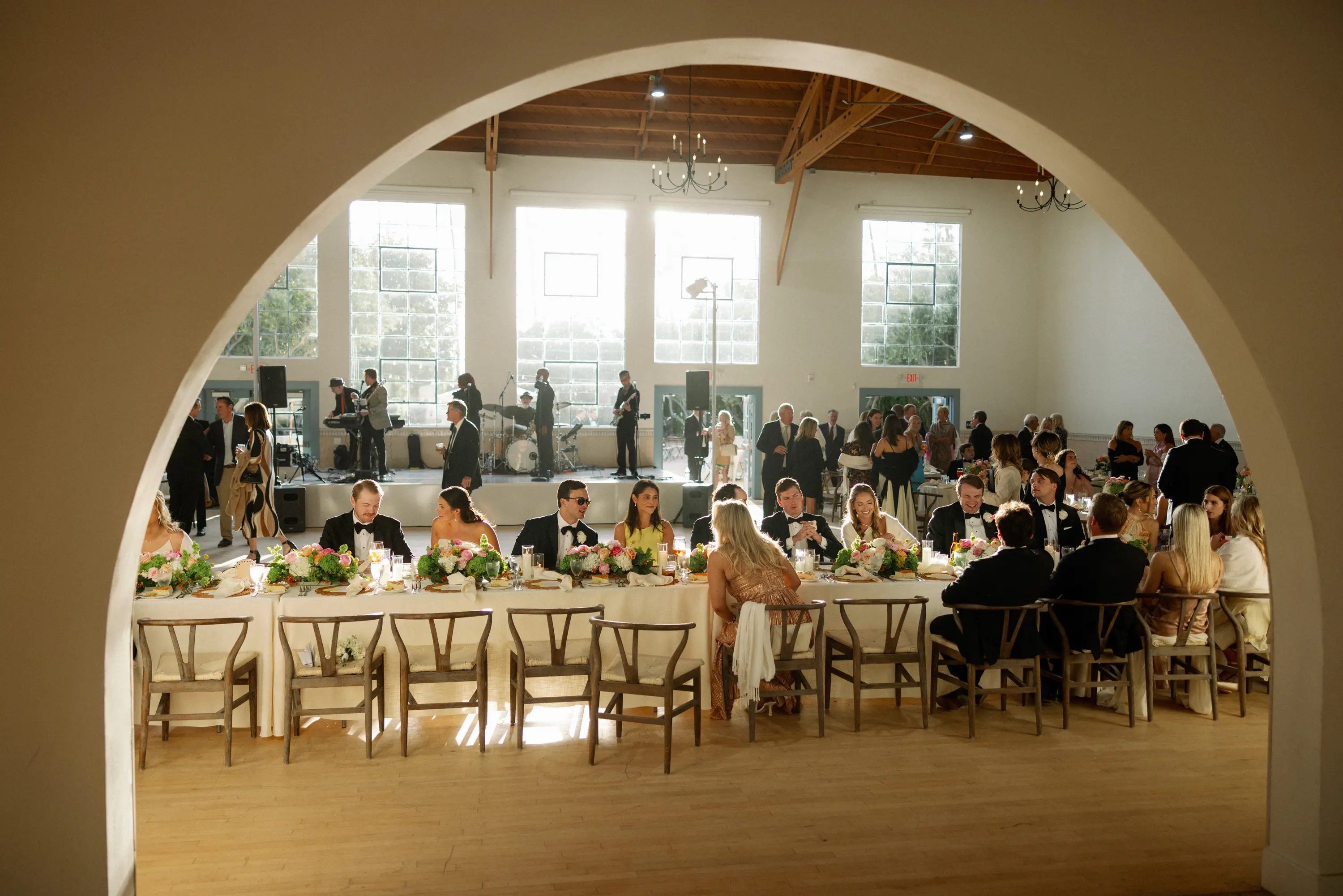 People in formal attire seated at a long banquet table with floral centerpieces inside a large, sunlit wedding venue.