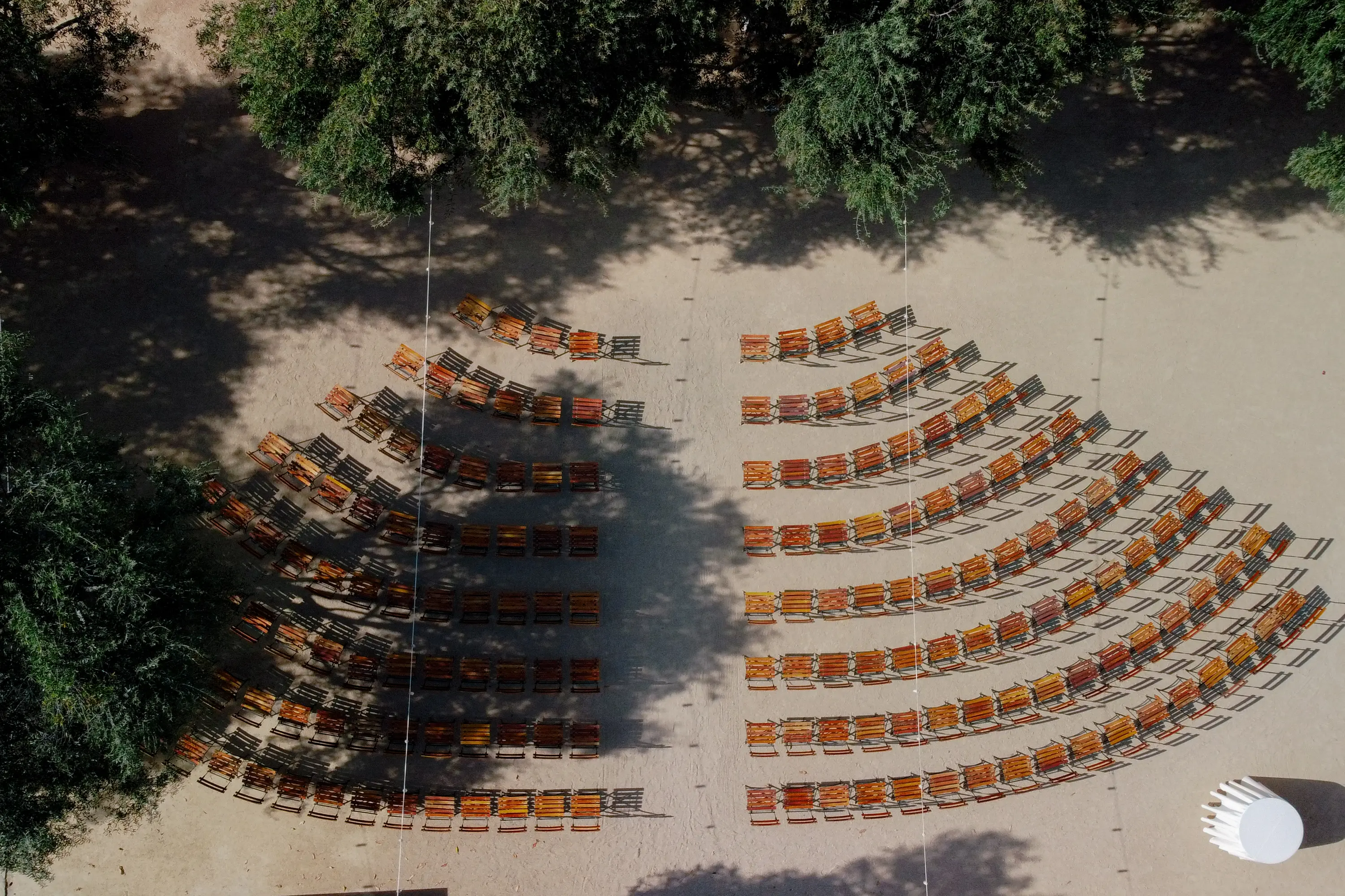 Aerial view of rows of empty orange chairs arranged outdoors in a semi-circle, with shadows cast by surrounding trees and a white covered round table in the bottom right.