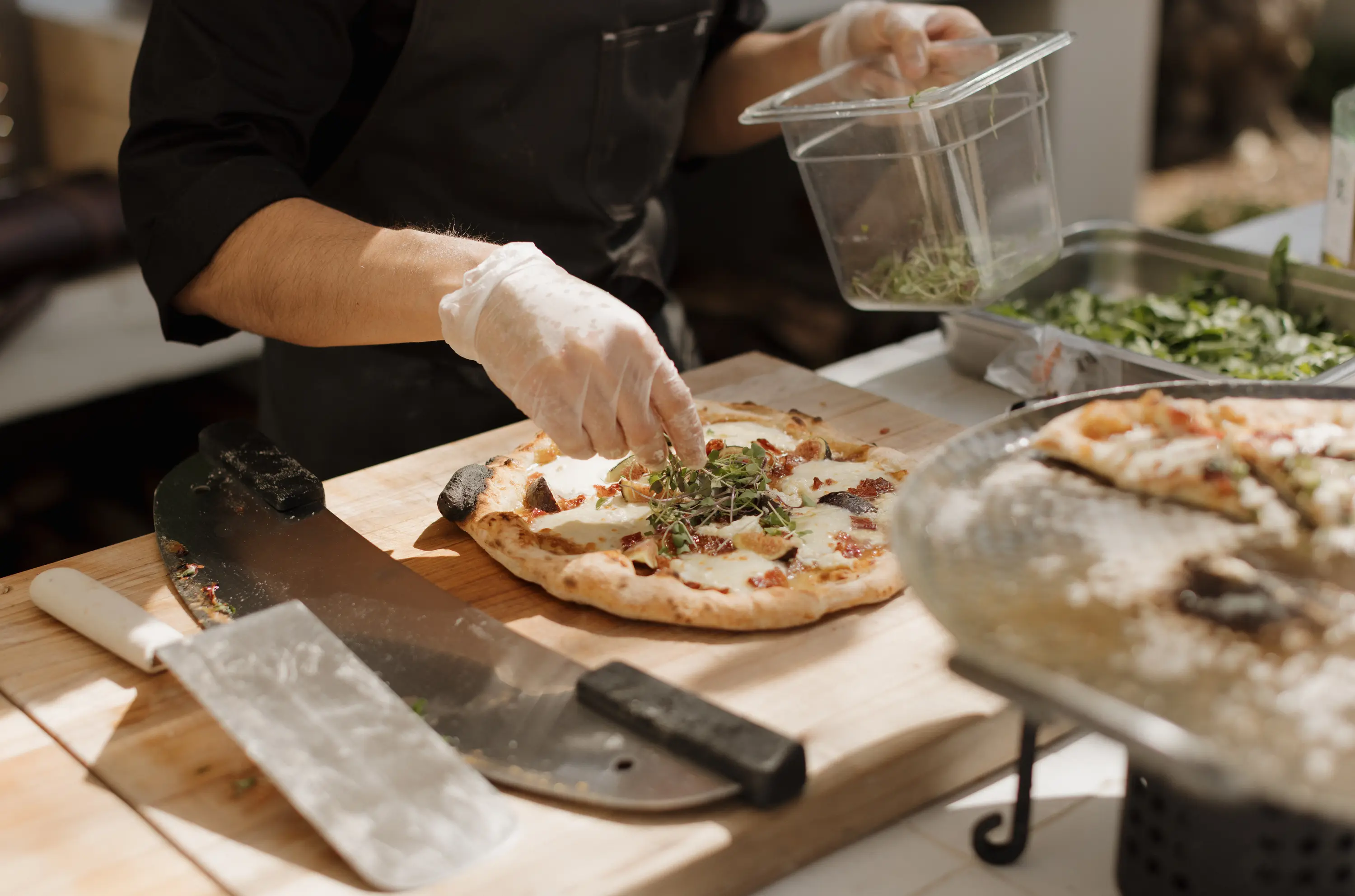 Chef wearing gloves garnishing a freshly baked pizza with greens on a wooden board next to pizza cutting tools.