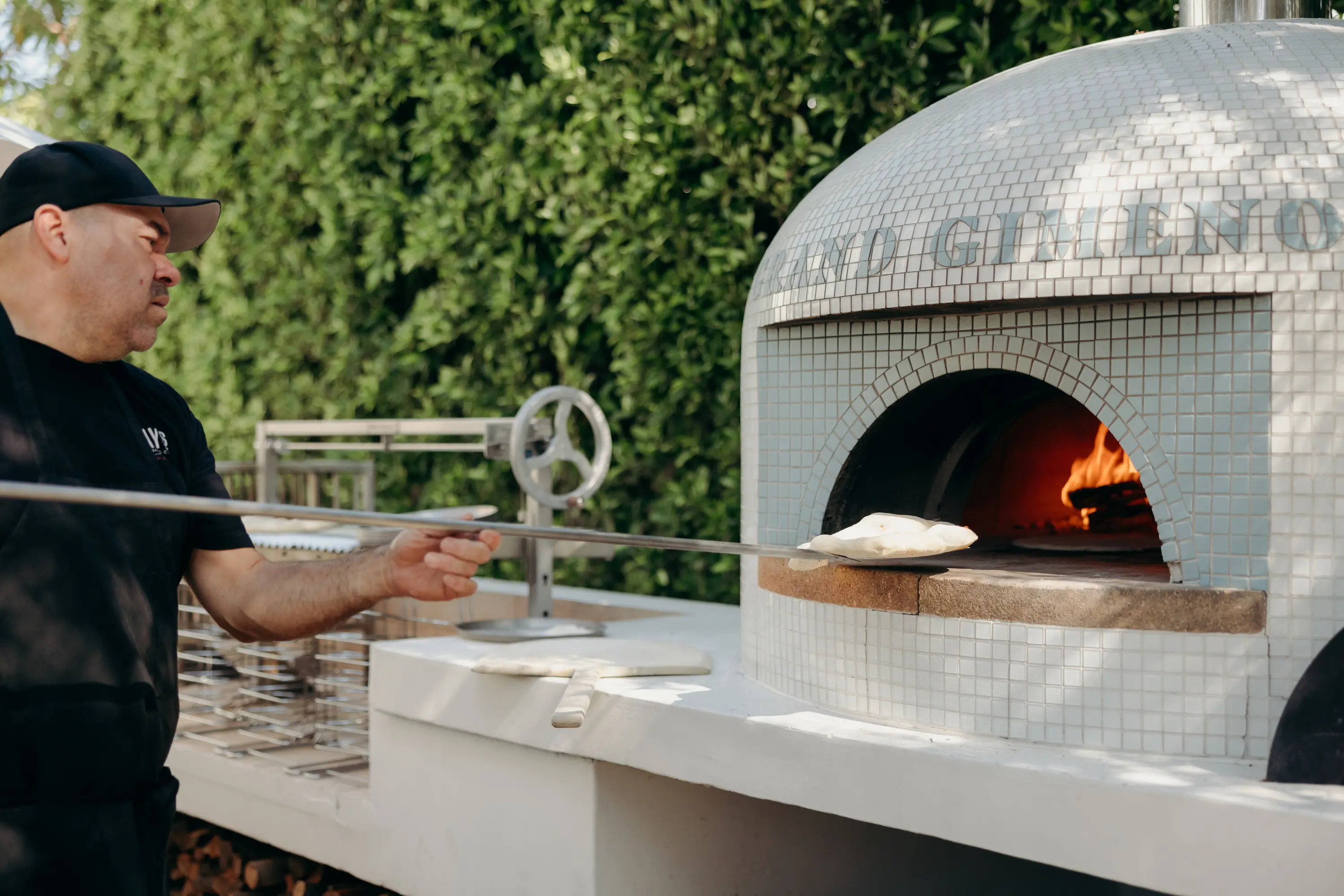 Man using a long-handled peel to place dough inside a tiled wood-fired pizza oven with a visible flame.