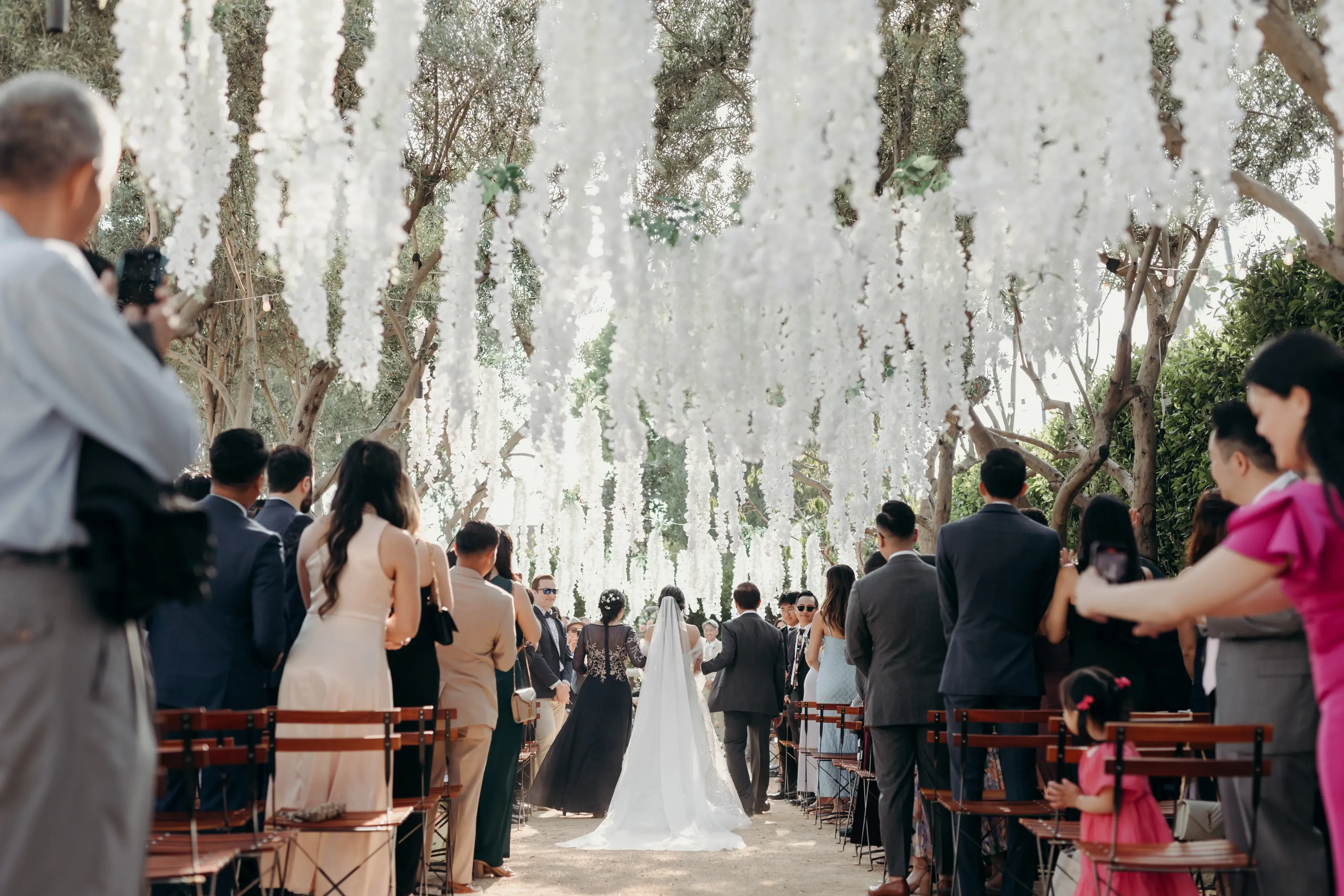Bride in white dress walking down outdoor aisle with guests standing on both sides under hanging white floral decorations.