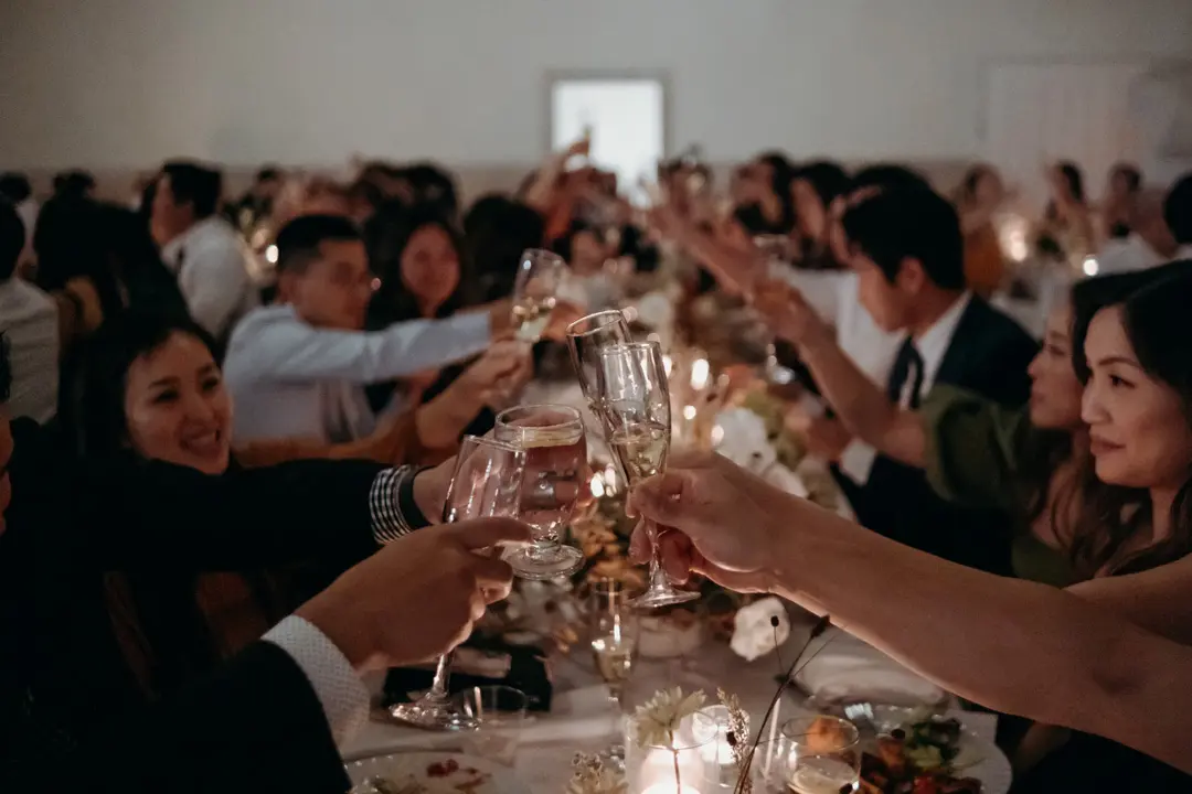 Guests raising glasses in a toast at a warmly lit dinner party or wedding reception.