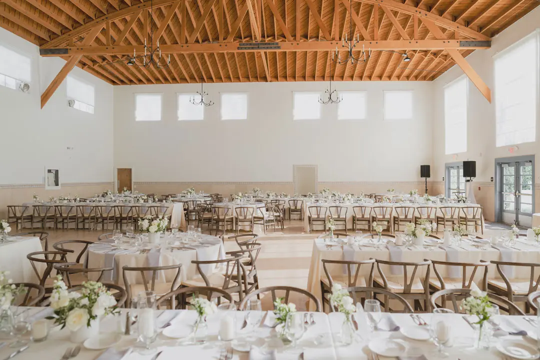 Bright, spacious wedding reception hall with wooden ceiling beams, round and rectangular tables decorated with white tablecloths and floral centerpieces.