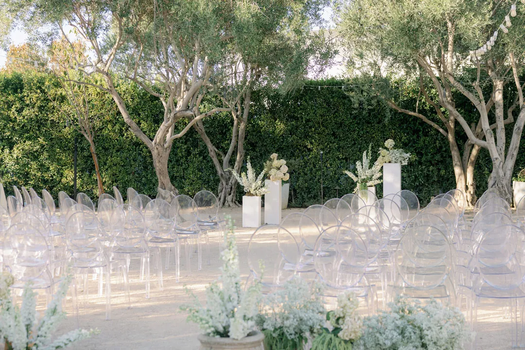 Clear ceremony chairs arranged along a flower-lined courtyard aisle