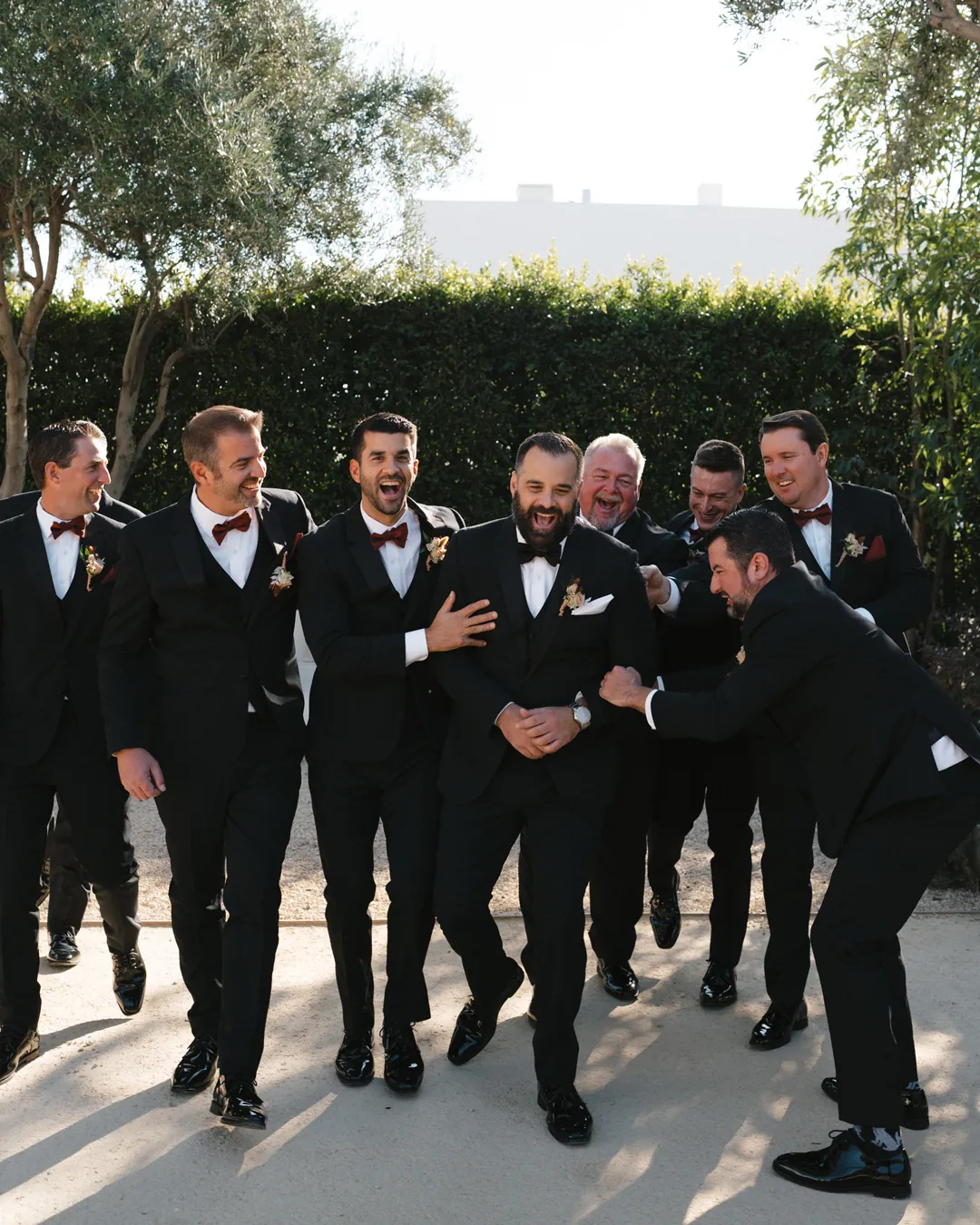 The groom and friends laugh together beneath olive trees.